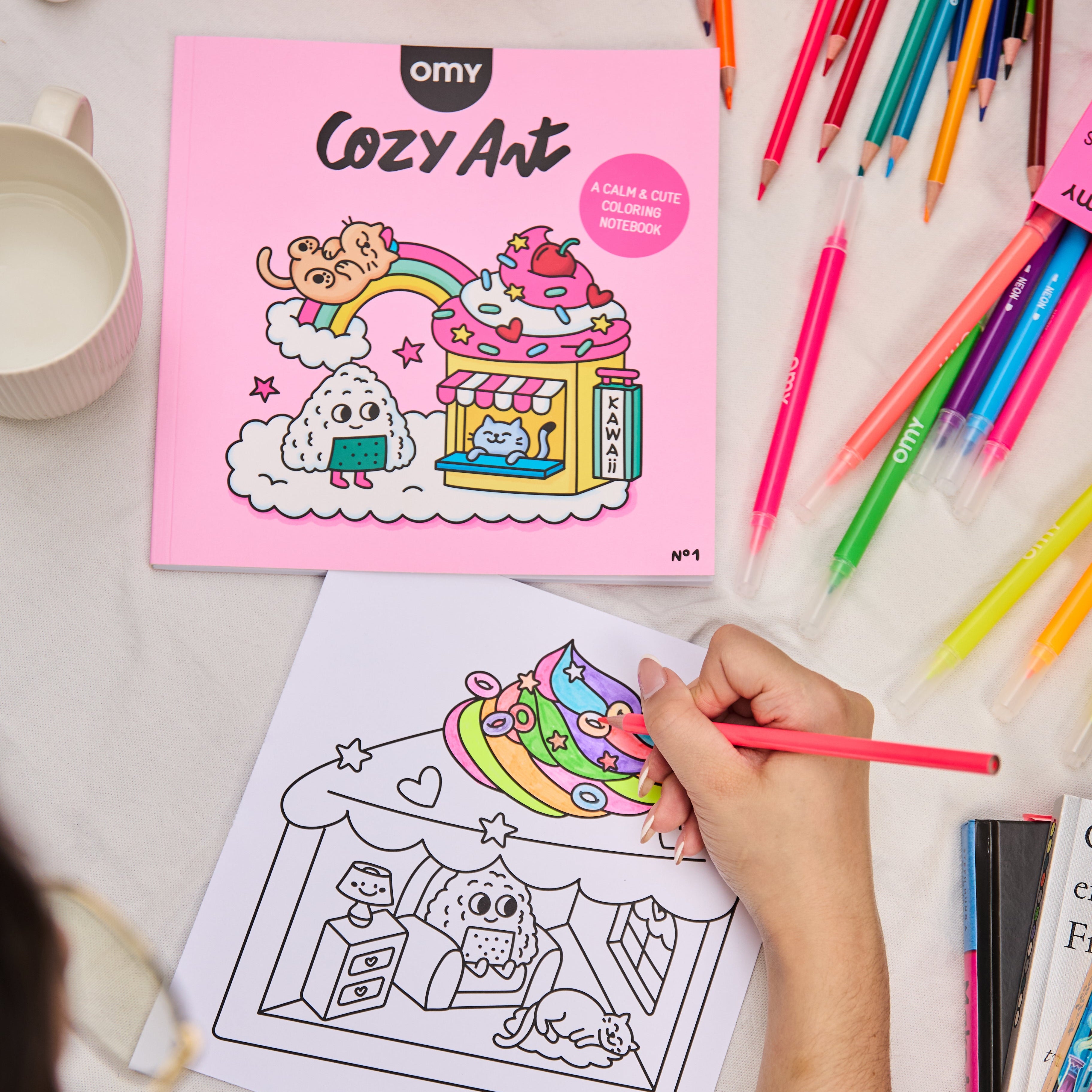 Person coloring a page from the Cozy Art Kawaii coloring book showing a cute dessert stand and rainbow, with colorful pencils scattered on the table, illustrating a relaxing and creative activity.