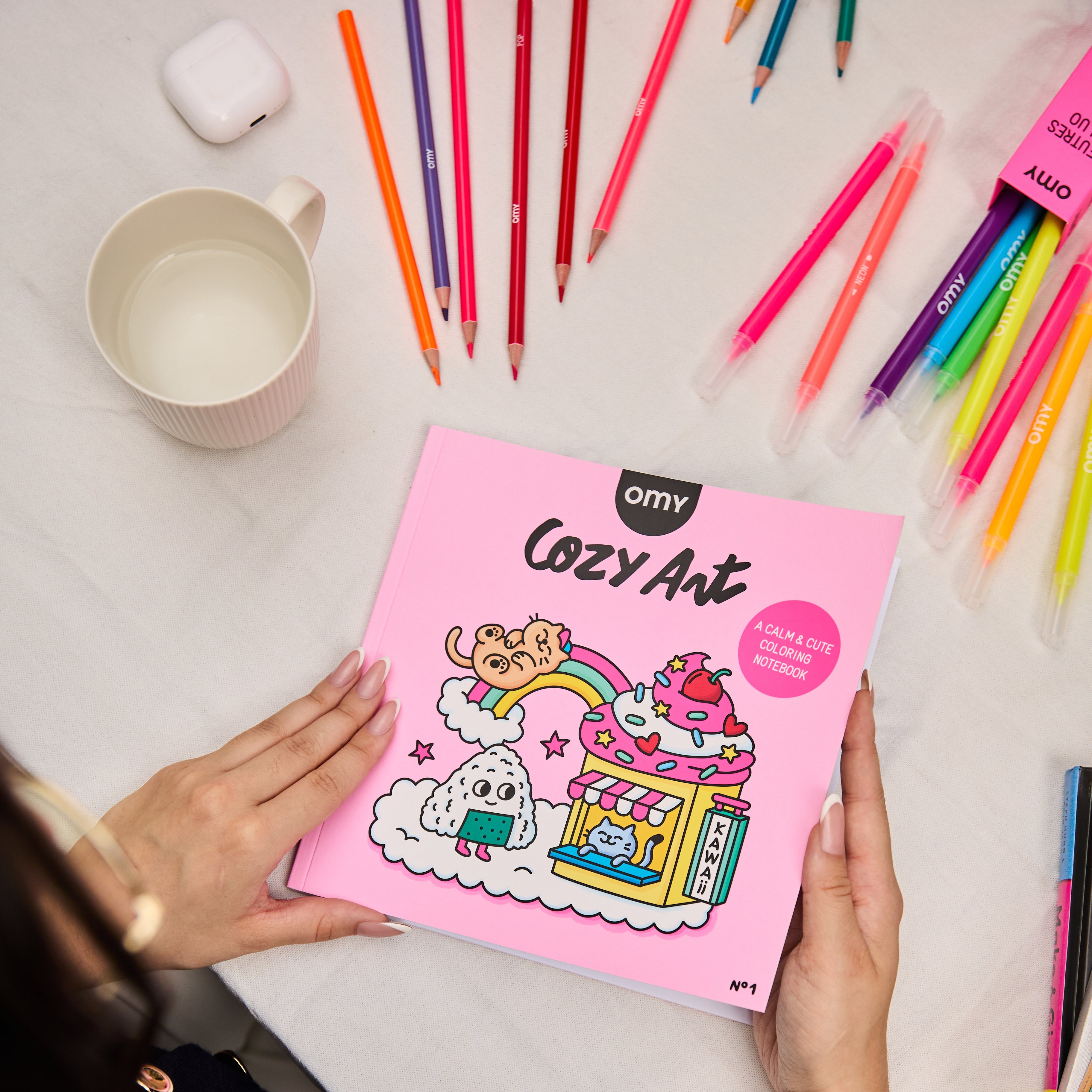 Hands holding the Cozy Art Kawaii coloring book closed with adorable kawaii characters like a bear sliding down a rainbow into an ice cream sundae, a rice ball, and a cat on the pink cover, surrounded by colorful pencils, markers, a cup and earbud case on the table.