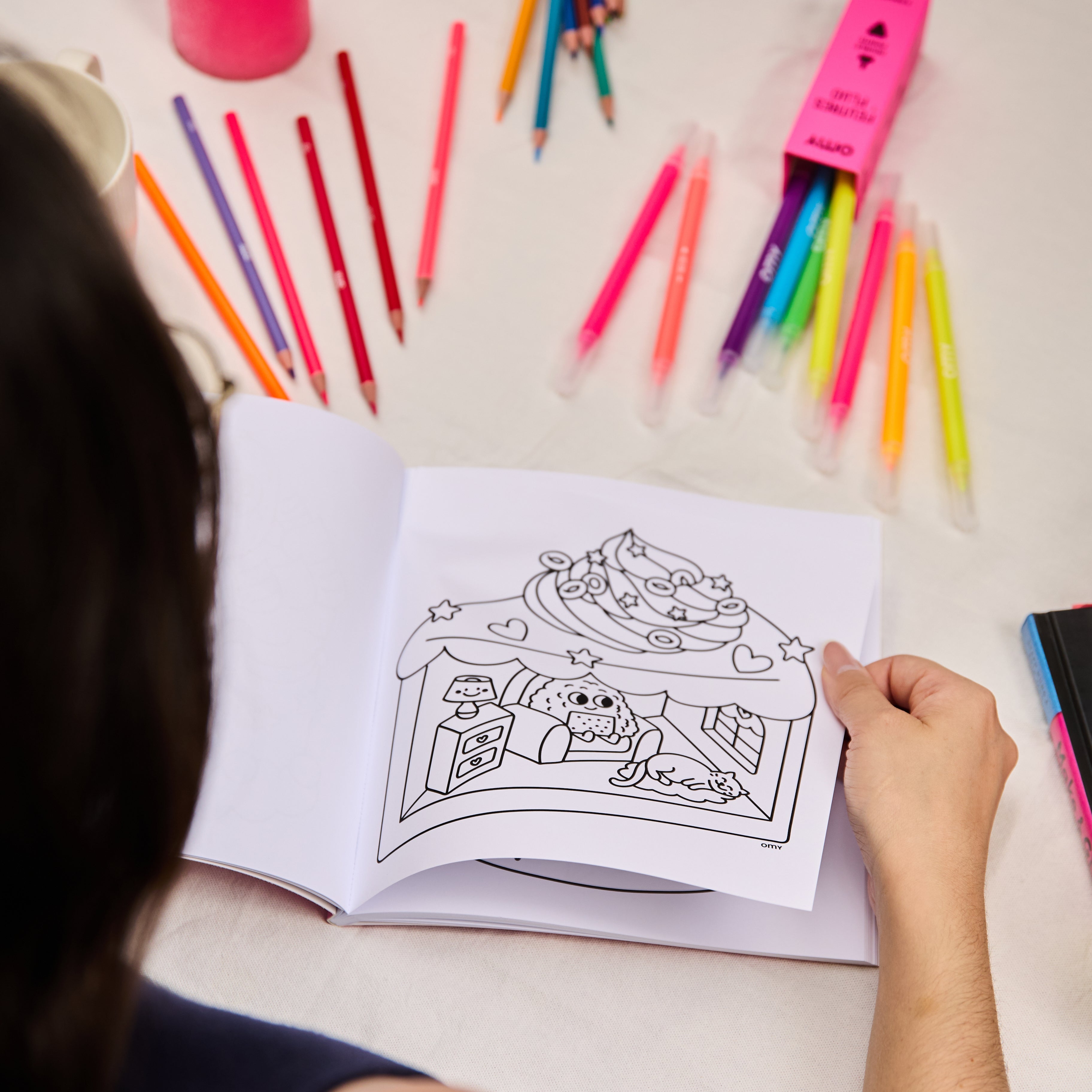 Open Cozy Art Kawaii coloring book showing a cute dessert shop illustration with hearts, with a person holding the page and colorful pencils and markers arranged on a table, perfect for creative coloring.