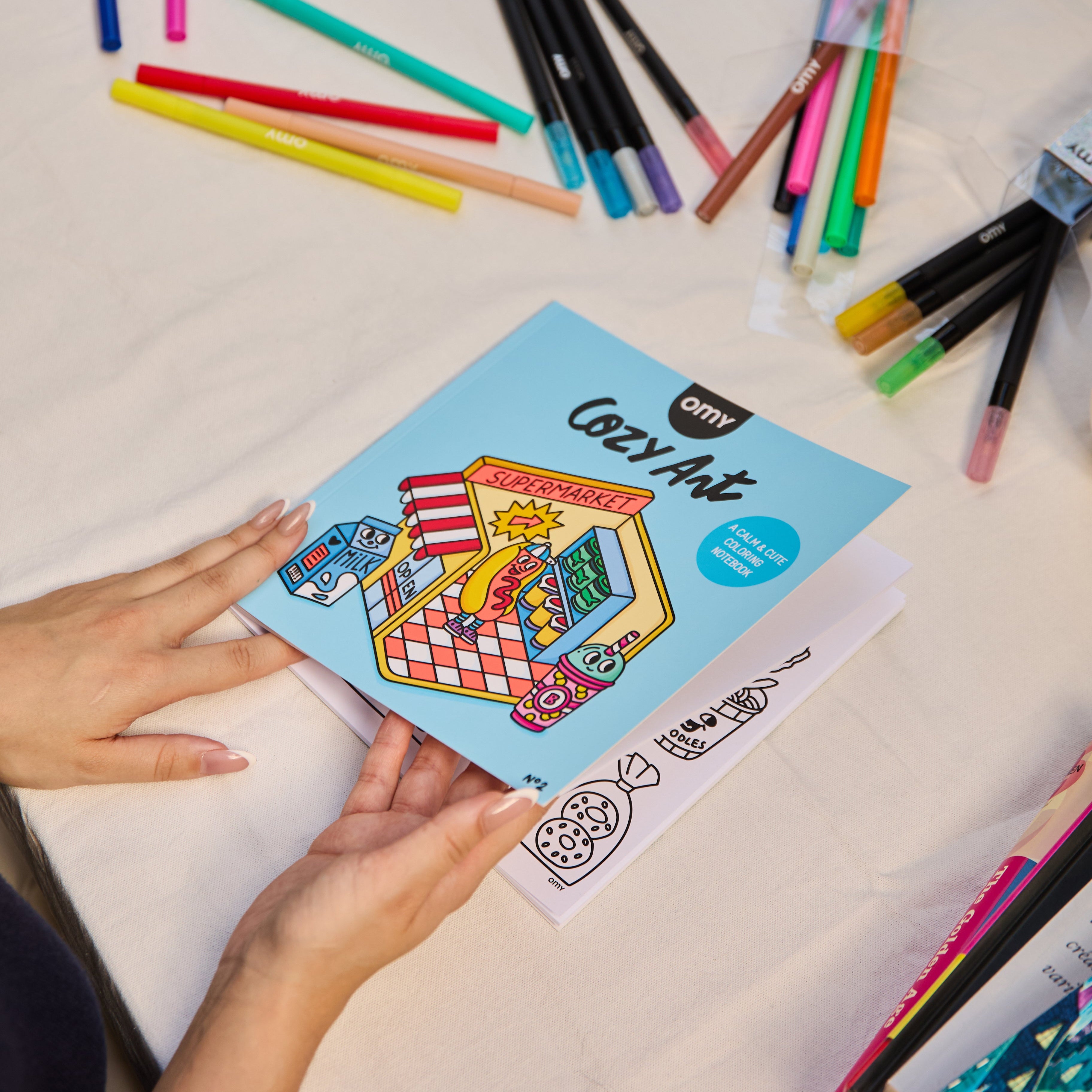 Hands holding the Cozy Art "Super Market" coloring notebook on a table surrounded by colorful markers