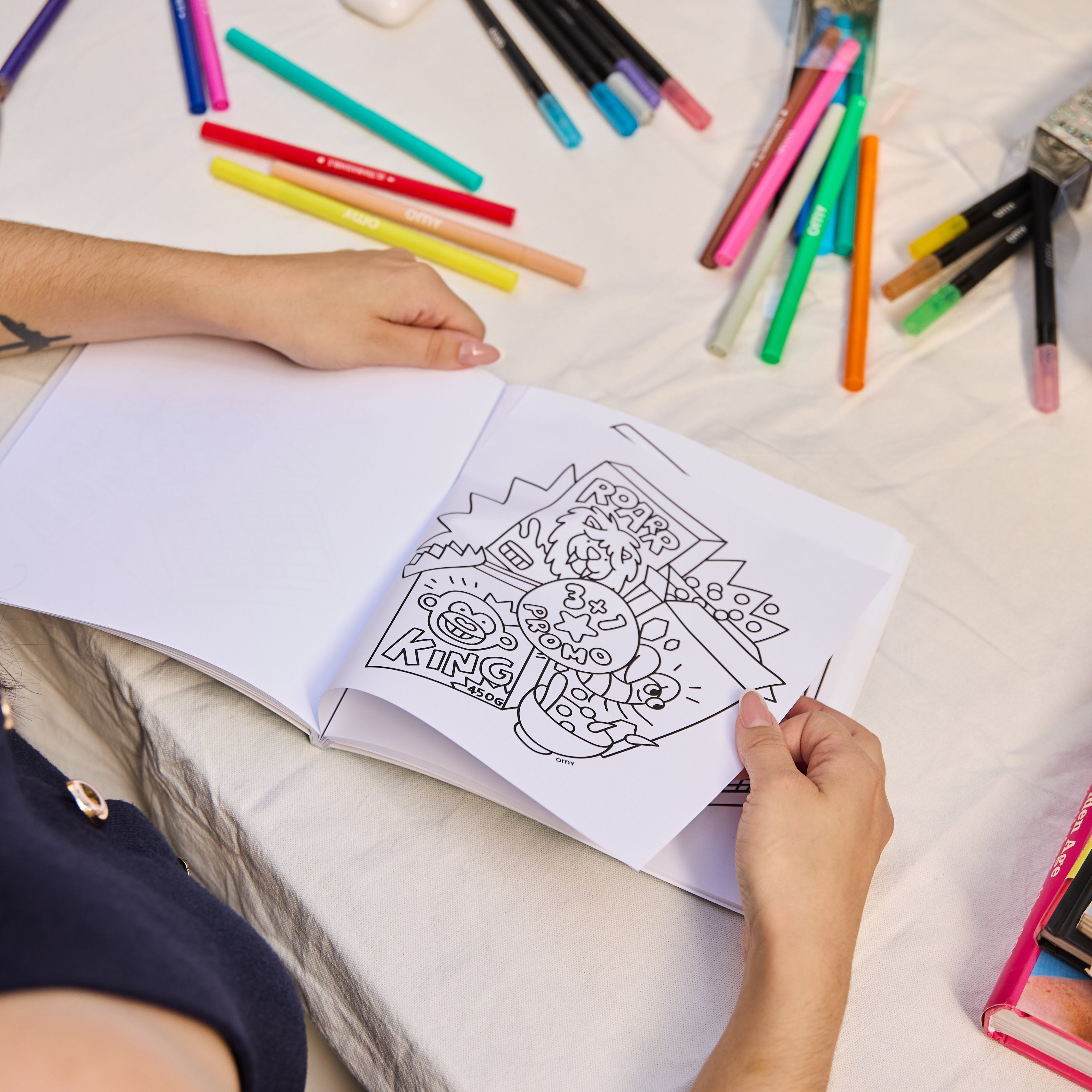 Person holding a coloring page from the Cozy Art "Super Market" notebook with a 'ROAR KING' design, with markers scattered on the table