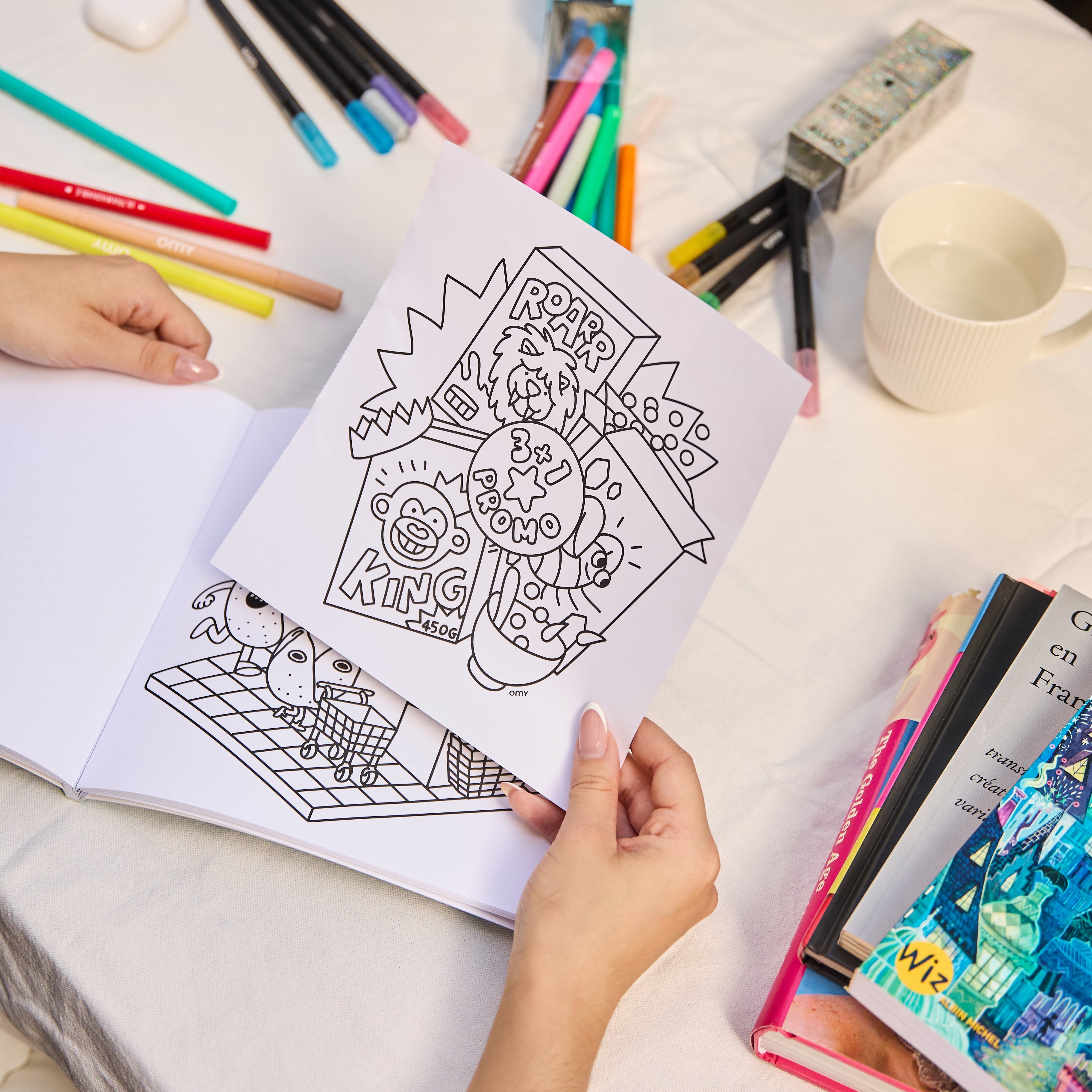 Hands tearing out a coloring page from the Cozy Art "Super Market" notebook with illustrated snack designs, surrounded by markers and a cup