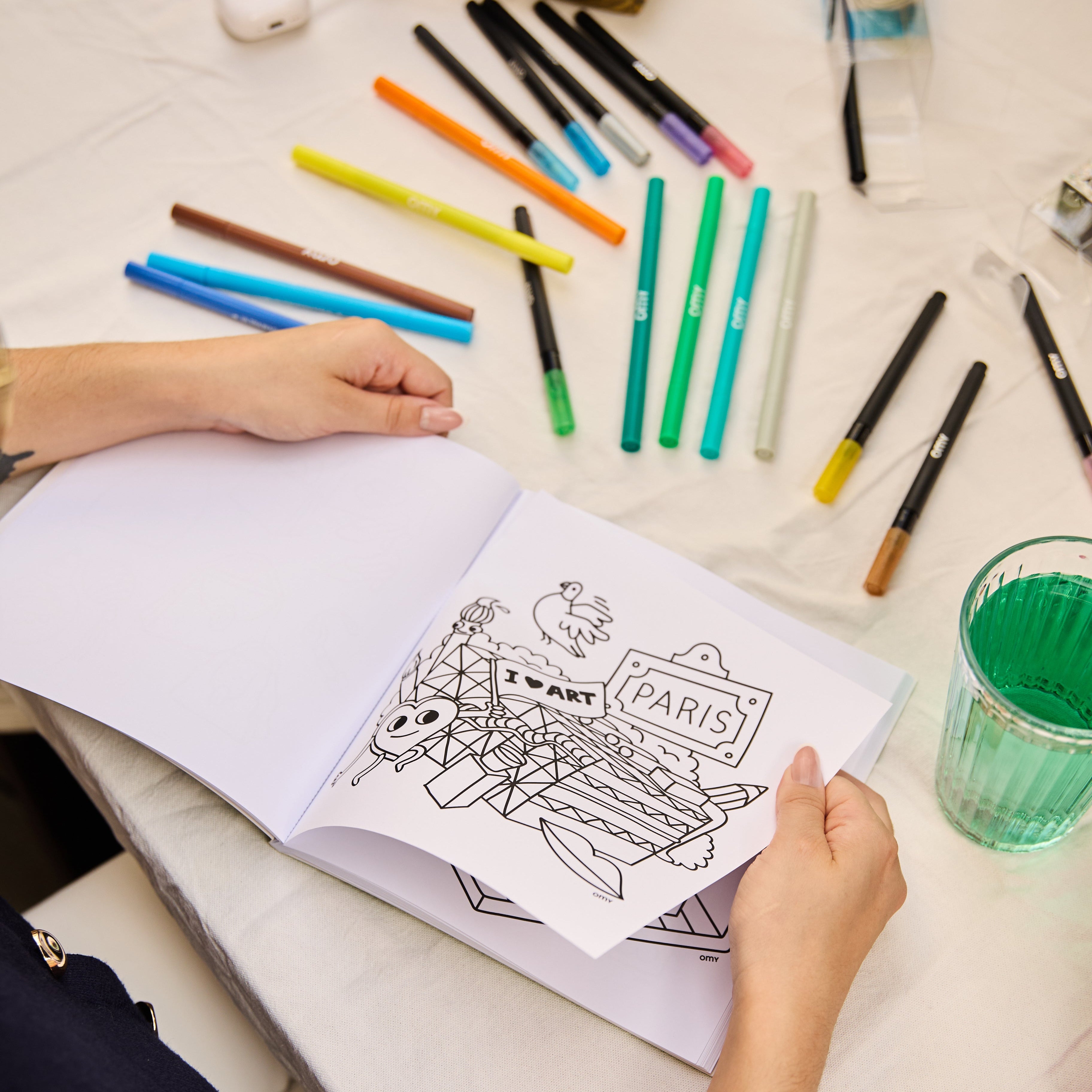 Hands holding the Cozy Art Paris coloring book with a Paris-themed coloring page partially cut out, surrounded by colorful markers on a table, illustrating creative crafting.
