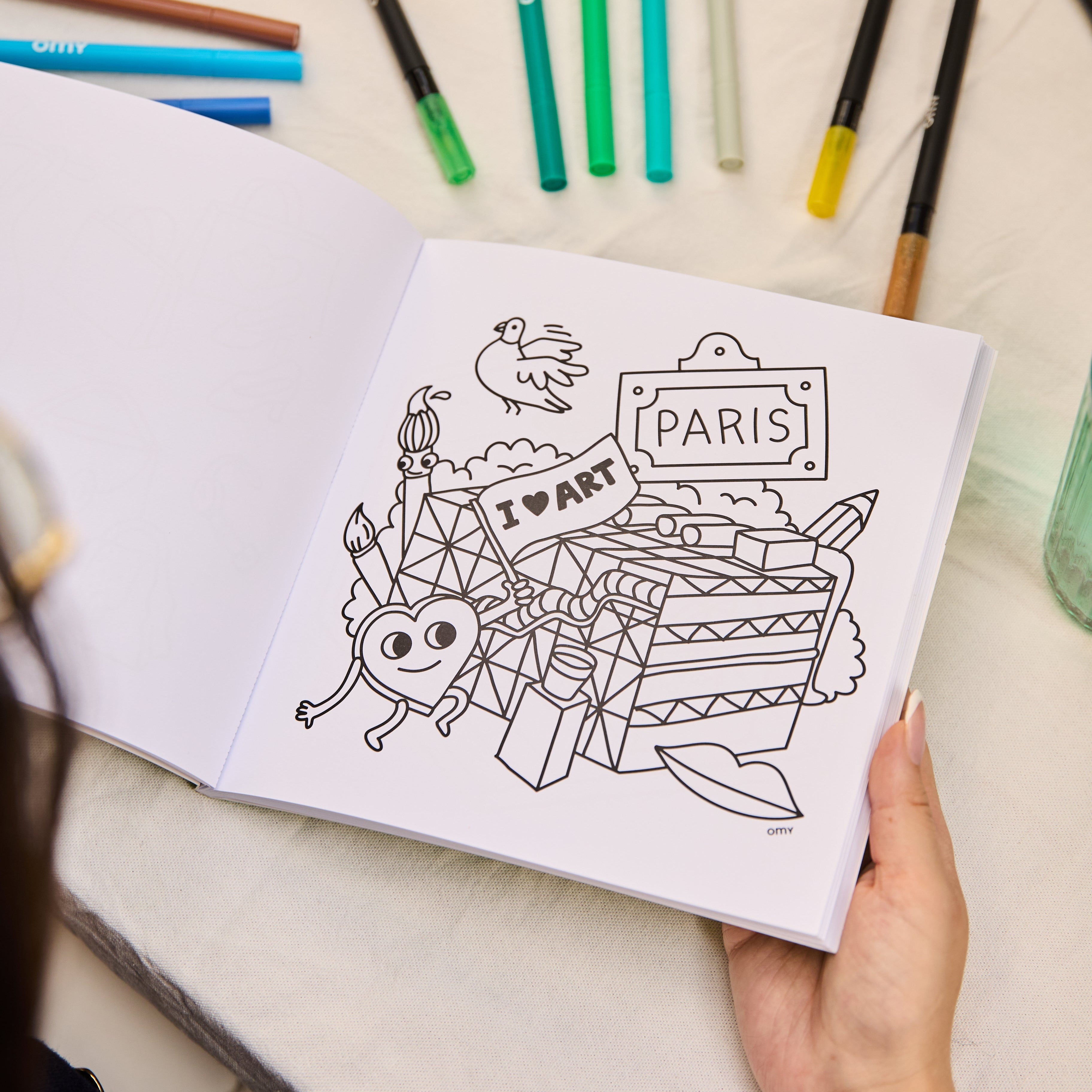 Open Cozy Art Paris coloring book displaying a coloring page with cute Paris-themed illustration featuring a Paris sign, smiling characters, and hearts, surrounded by colorful markers on a table.