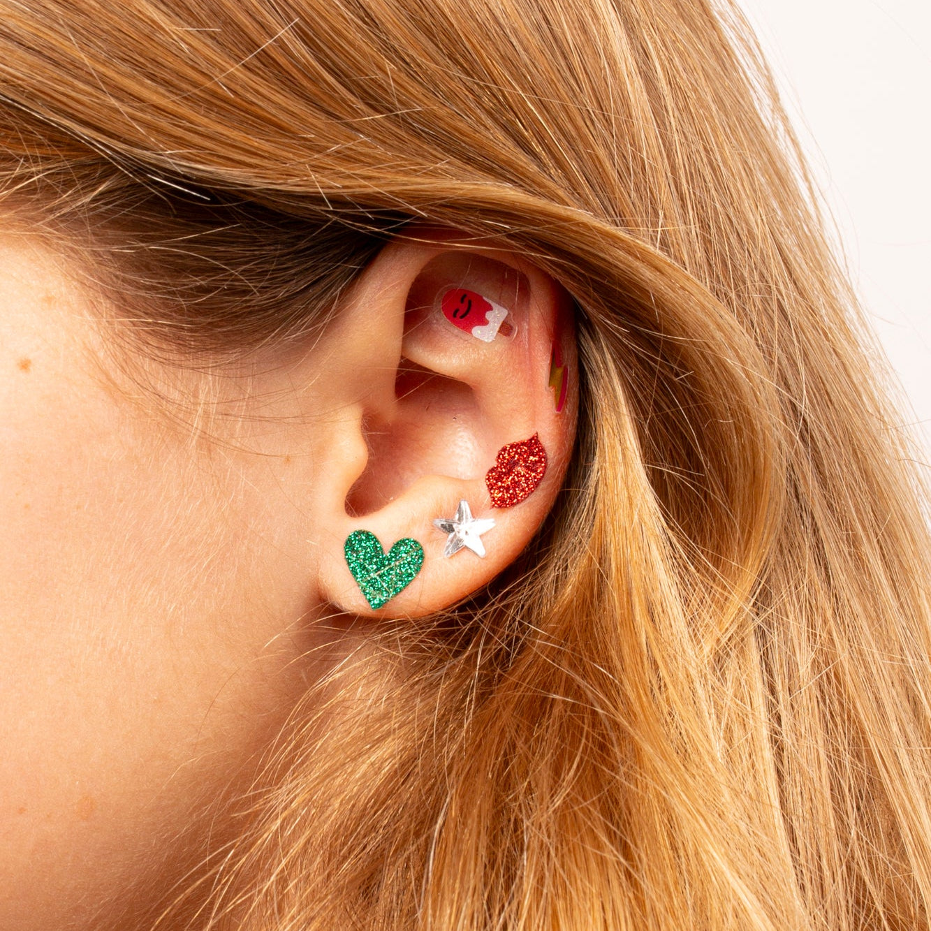 Close-up of an ear wearing colorful heart-shaped earrings Kawai OMY with blonde hair.