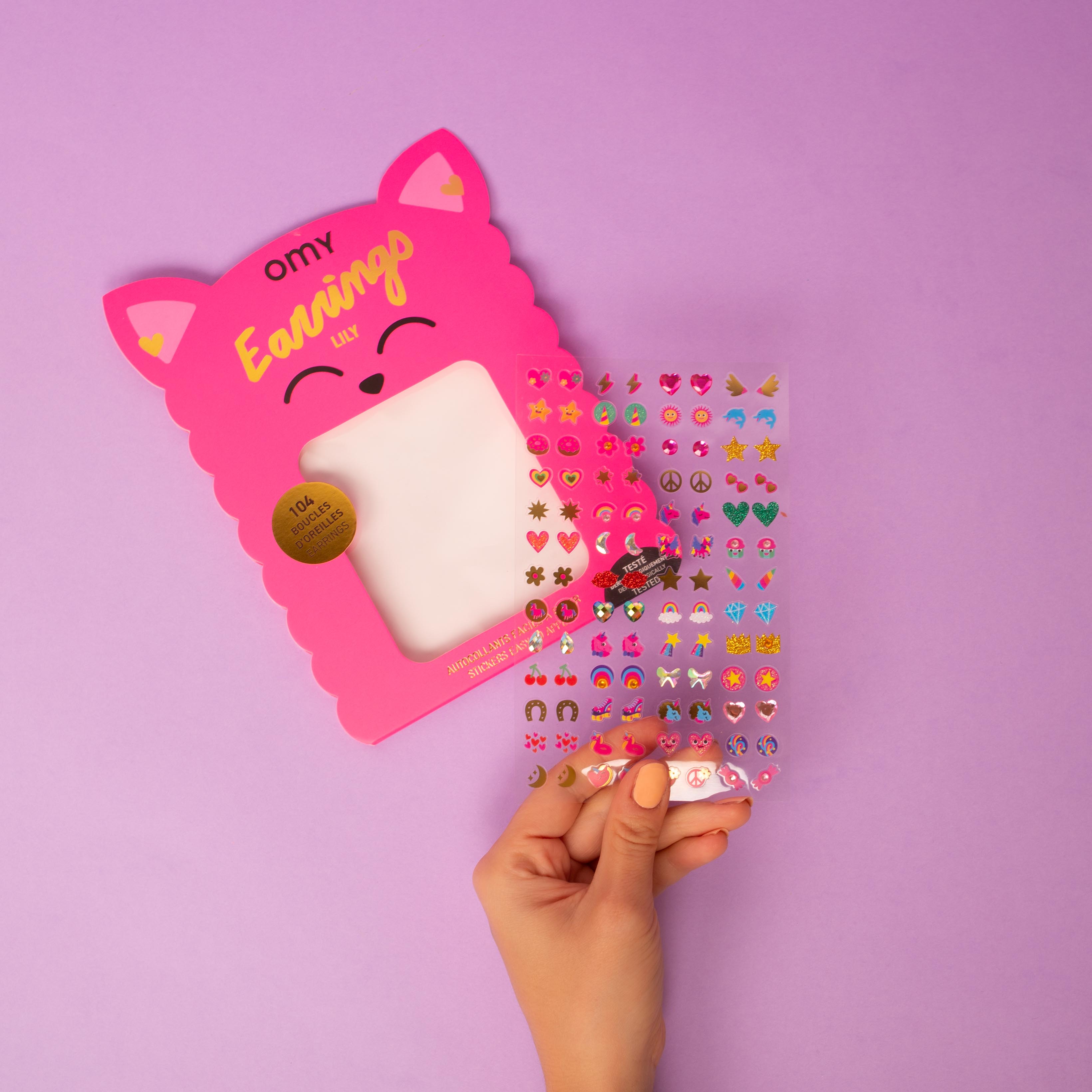 Hand holding a pink package of earrings Lily OMY with a purple background