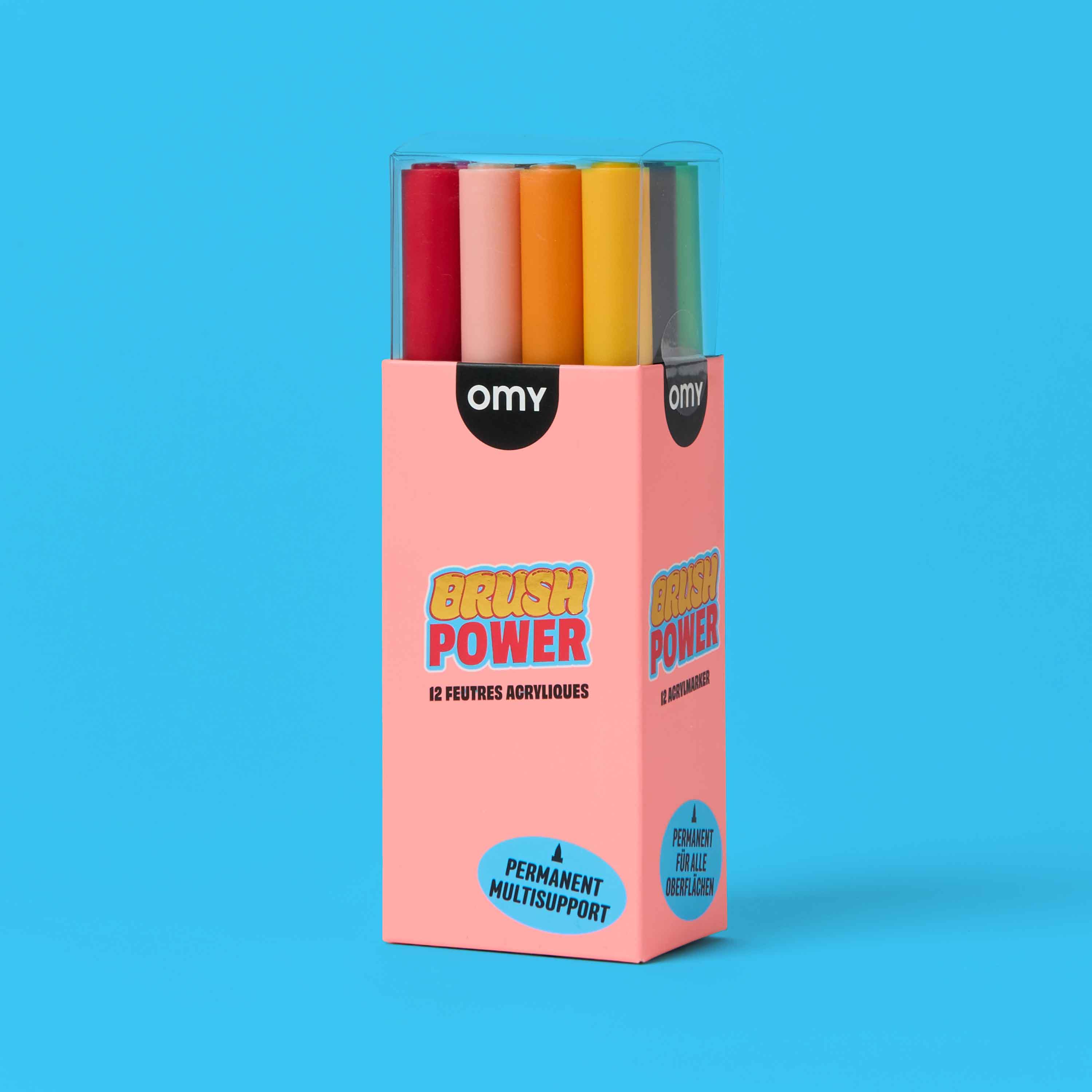 OMY Brush Power 12 acrylic markers in pink open packaging – permanent multi-surface brush tip pens with colorful caps including red, orange, yellow and green on blue background