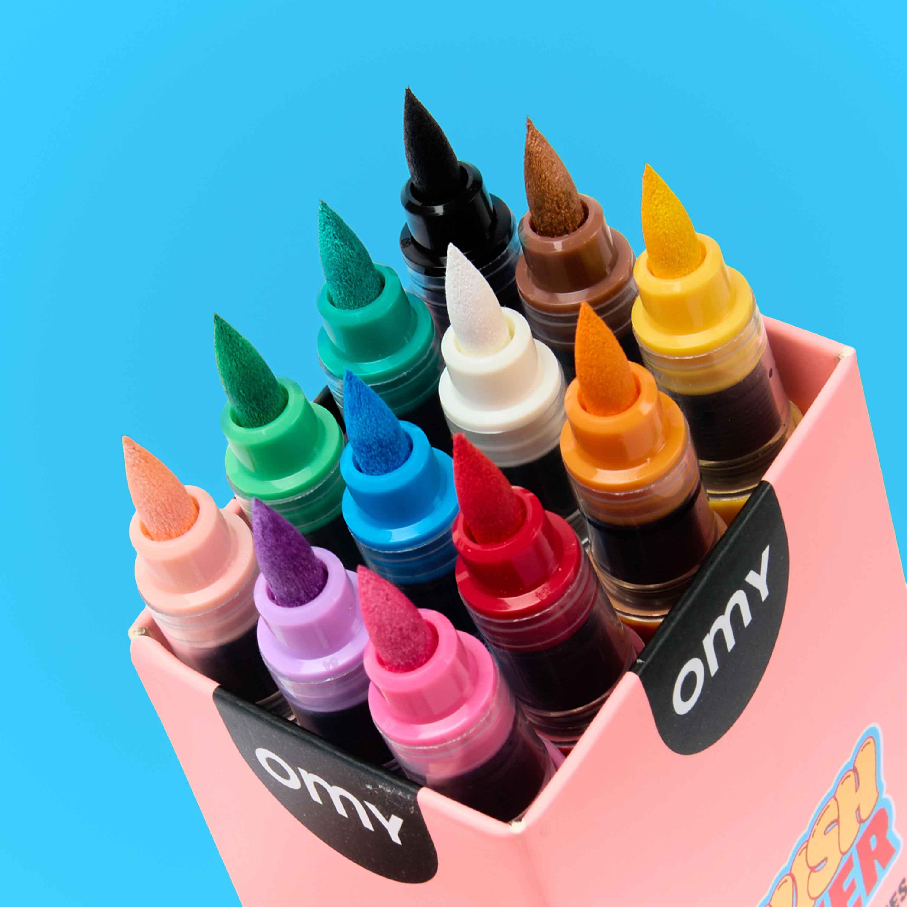 Top view of OMY Brush Power marker set in a pink box – colorful brush tip markers including green, blue, pink, purple, white, black, orange, brown and yellow on blue background