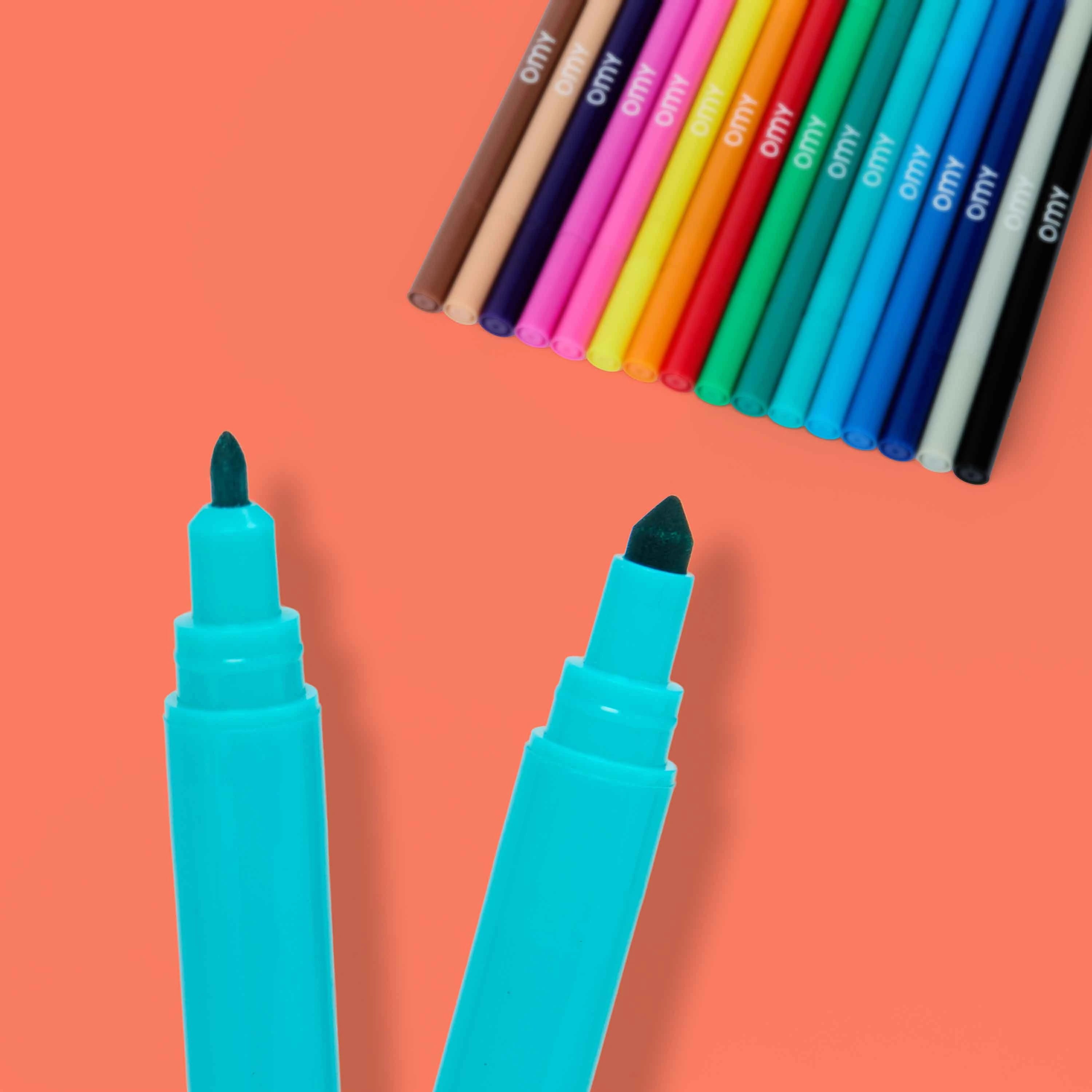 OMY Super Clean 16 washable dual-tip markers creative flat lay – two teal brush tip markers in foreground with full rainbow set fanned out on coral background