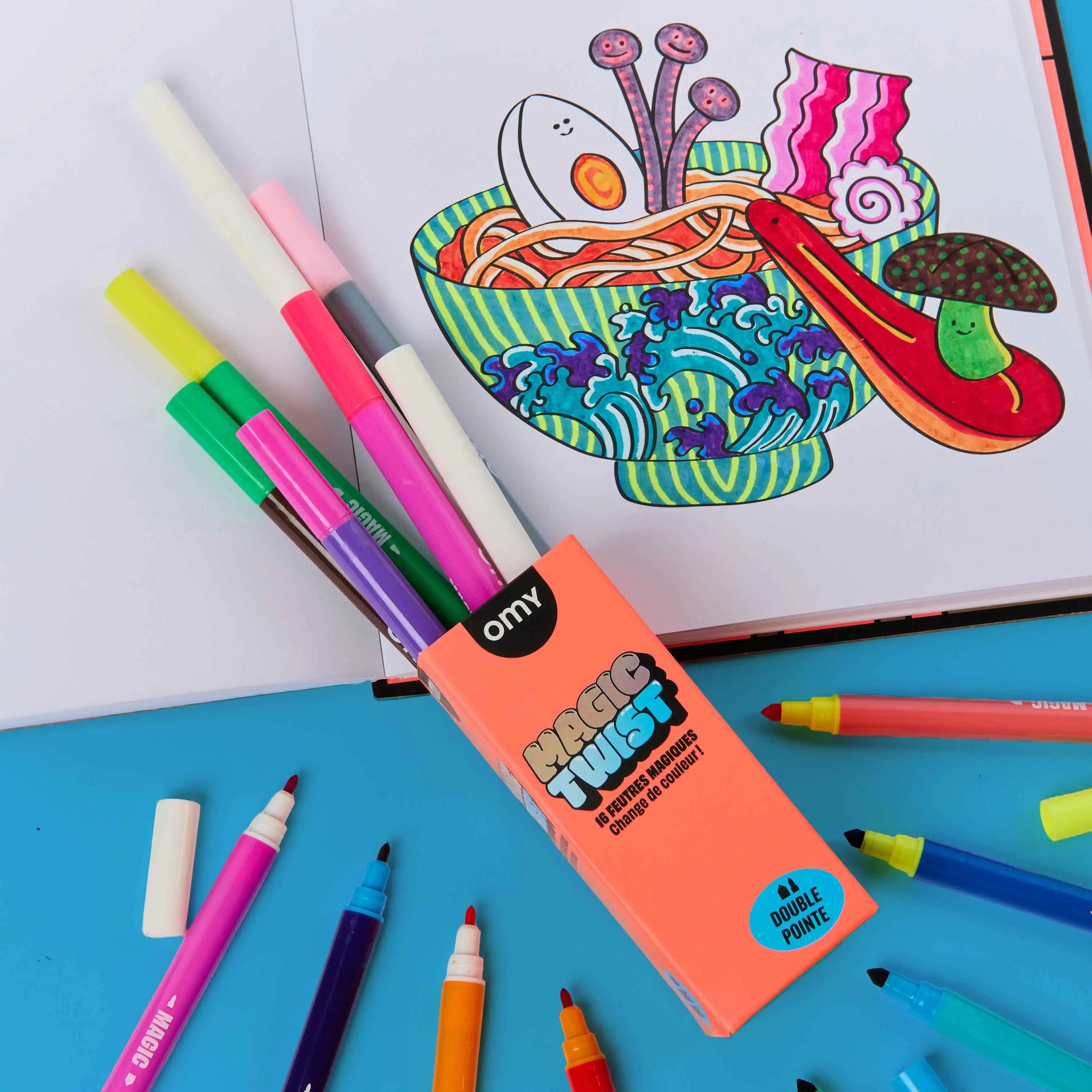 OMY Magic Twist 16 color-changing dual-tip markers in coral packaging surrounded by colorful felt tip pens on blue background, with a completed kawaii ramen bowl coloring page