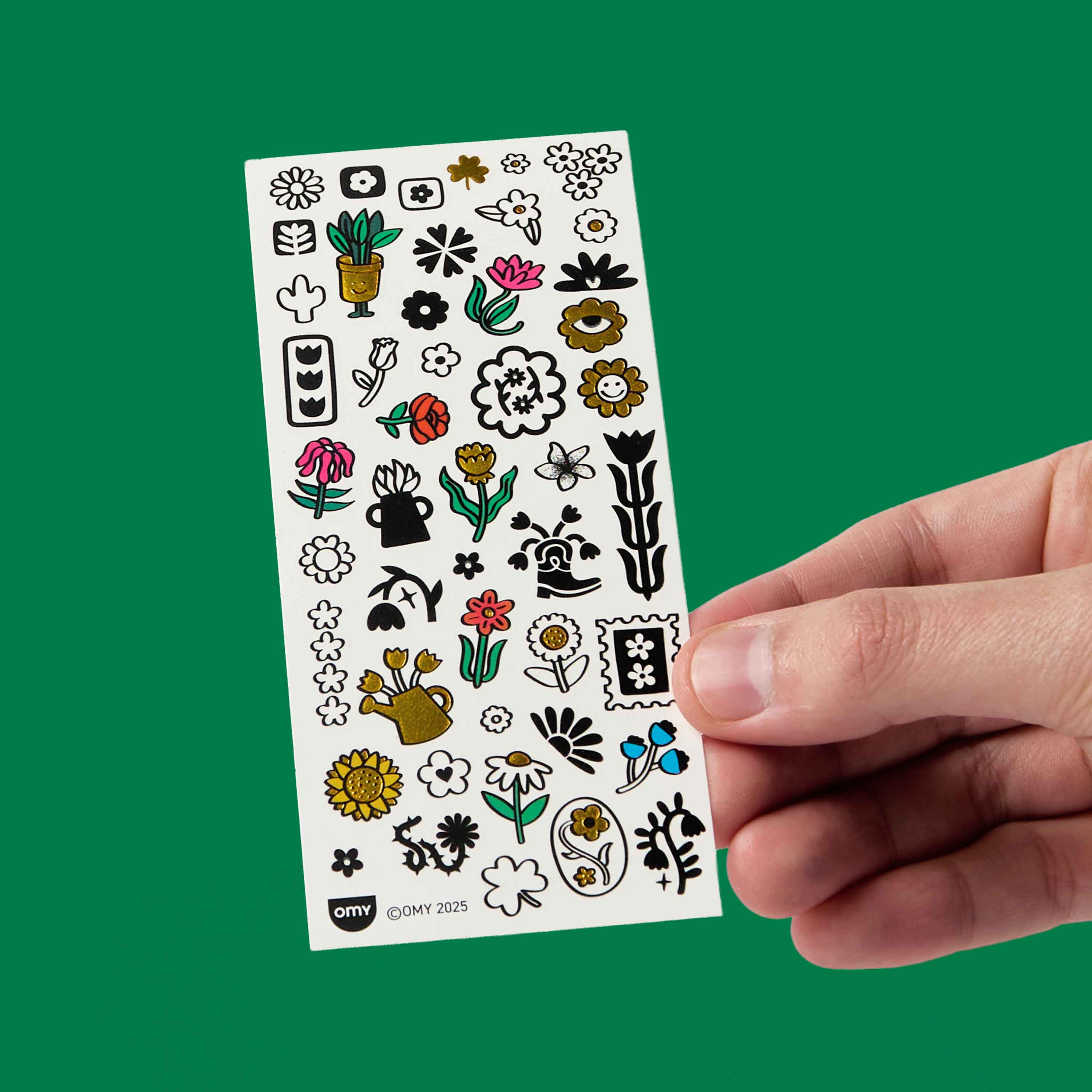 Sheet of floral and plant-themed stickers flash tattoos OMY held by a hand against a green background