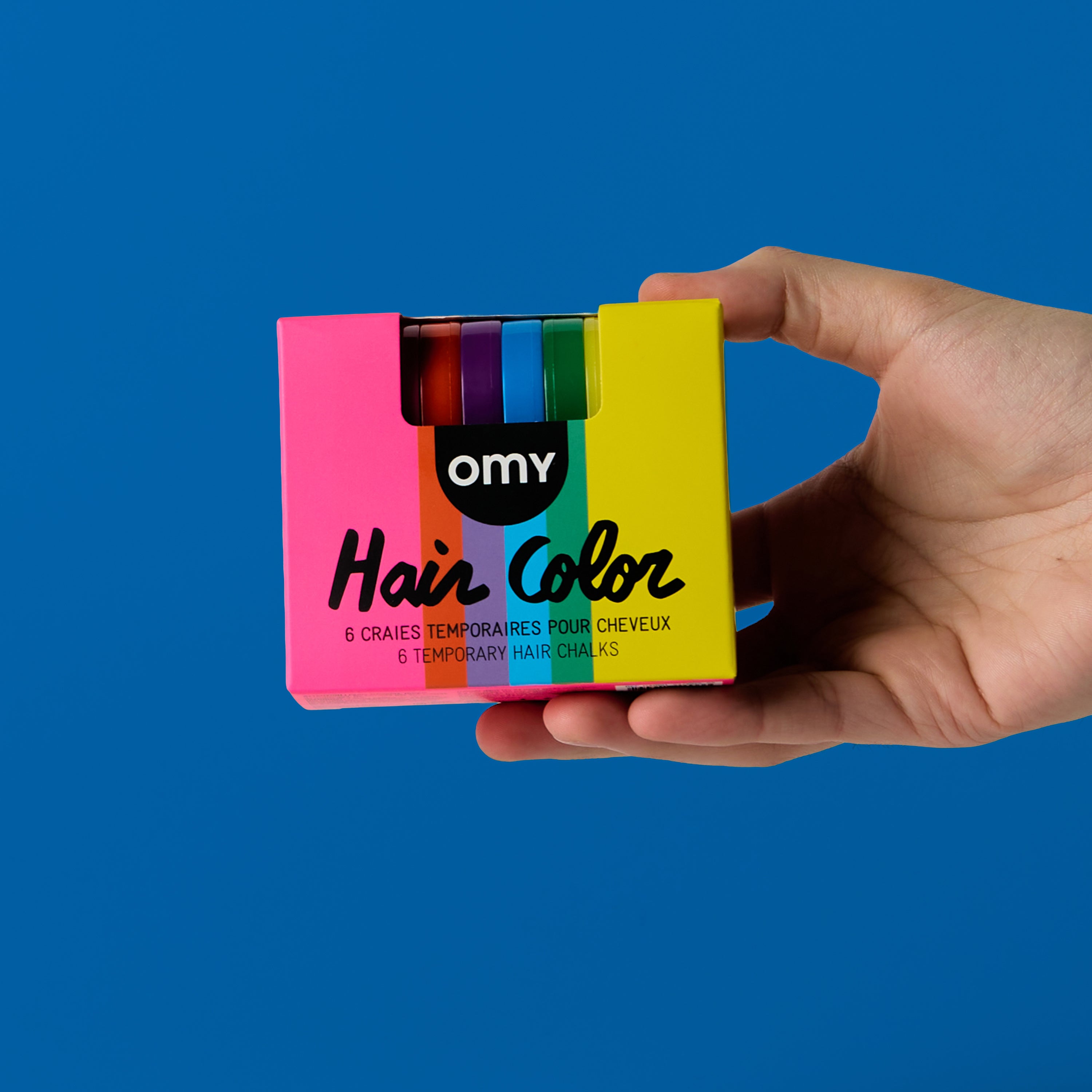 Hand holding OMY Hair Color Rainbow box β 6 temporary hair chalks in rainbow colors including pink, yellow, green, blue, red and purple on blue background