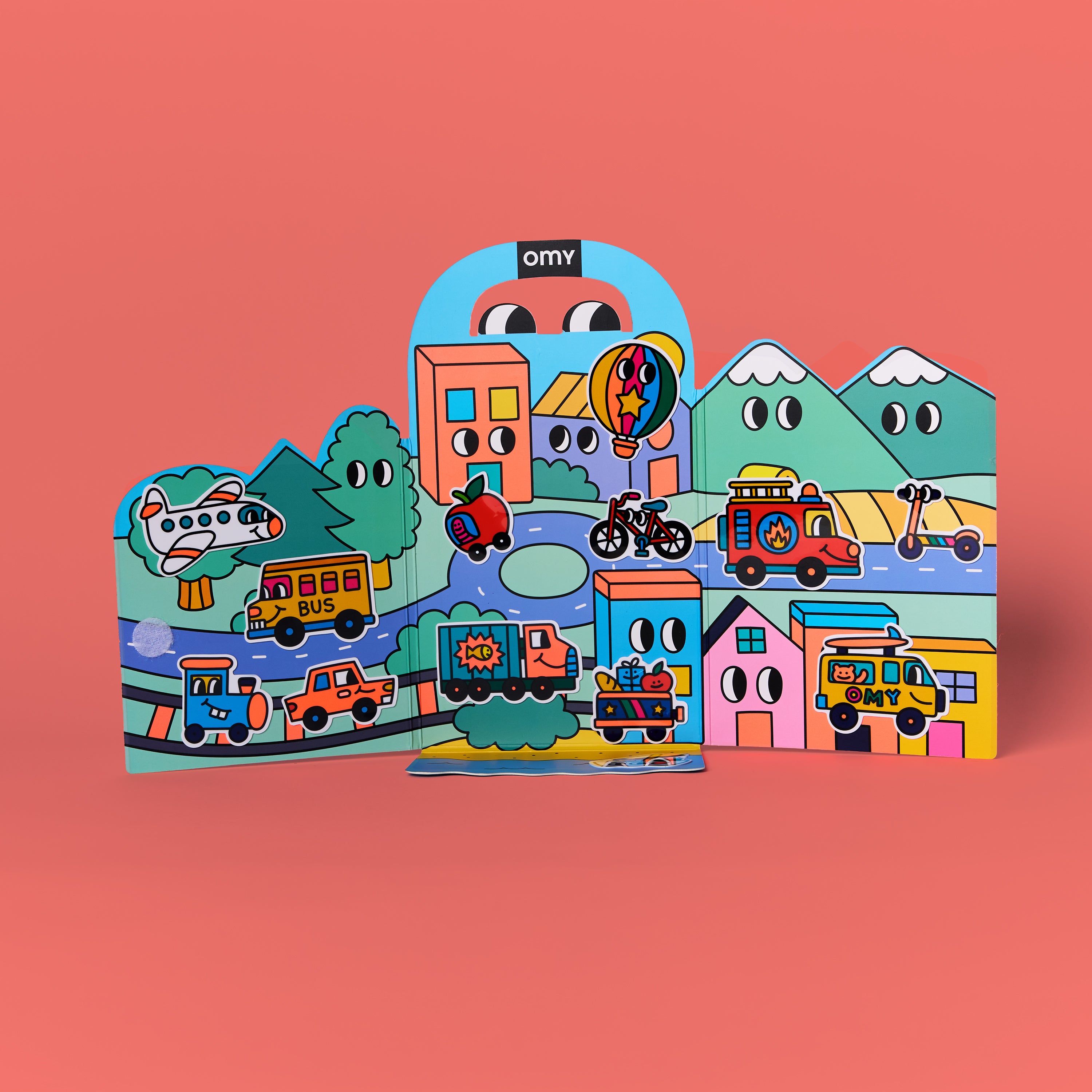 Fold-out scene from OMY Montessori Stickers Kit – Vroom Vroom edition standing upright on coral background featuring colorful town, mountains, roads and vehicles with reusable silicone stickers; creative play for kids aged 3–7