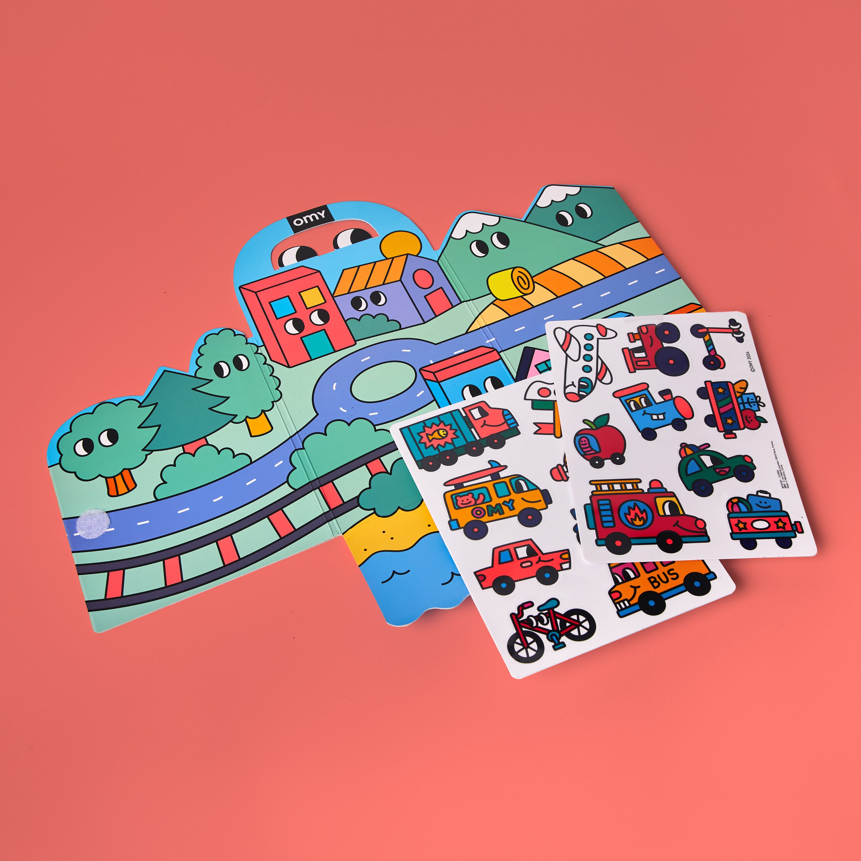 Fold-out scene and reusable silicone sticker sheets from OMY Montessori Stickers Kit – Vroom Vroom edition on coral background, featuring colorful town, roads and vehicles like bus, fire truck, train and helicopter; repositionable travel activity for kids aged 3–7