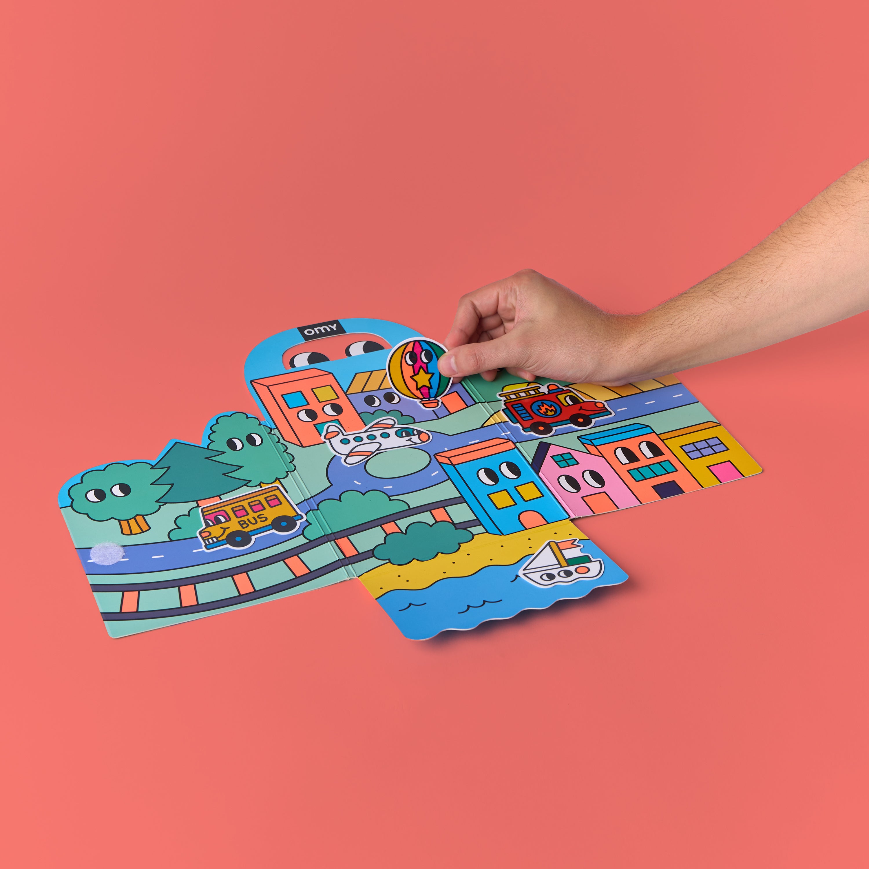 Hand placing reusable silicone sticker onto fold-out scene from OMY Montessori Stickers Kit – Vroom Vroom edition, showing colorful town, train tracks, roads and houses on coral background; repositionable sticker activity for kids aged 3–7
