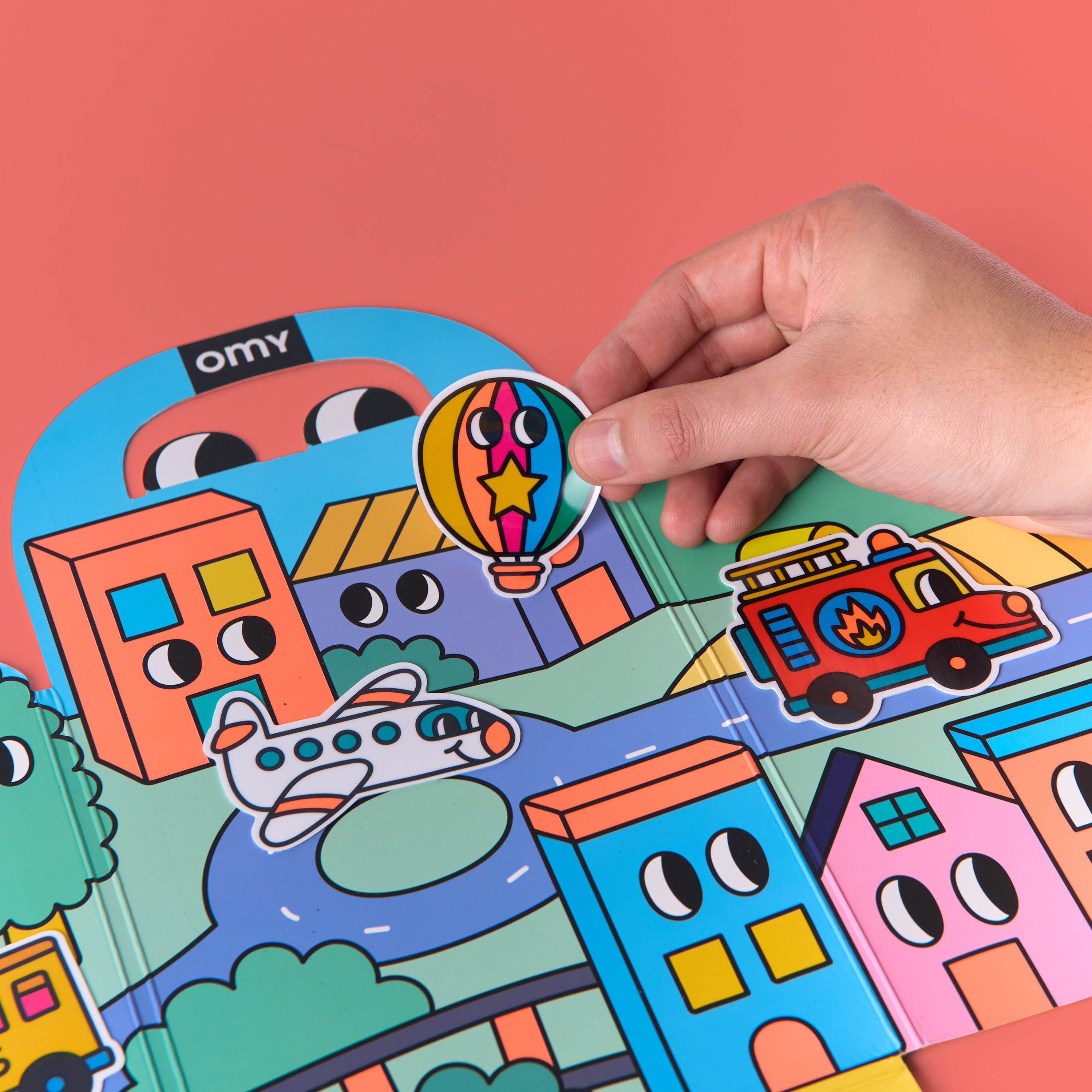 Close-up of hand placing hot air balloon sticker onto fold-out scene from OMY Montessori Stickers Kit – Vroom Vroom edition, featuring colorful buildings, airplane, fire truck and roads on coral background; creative screen-free play for kids aged 3–7