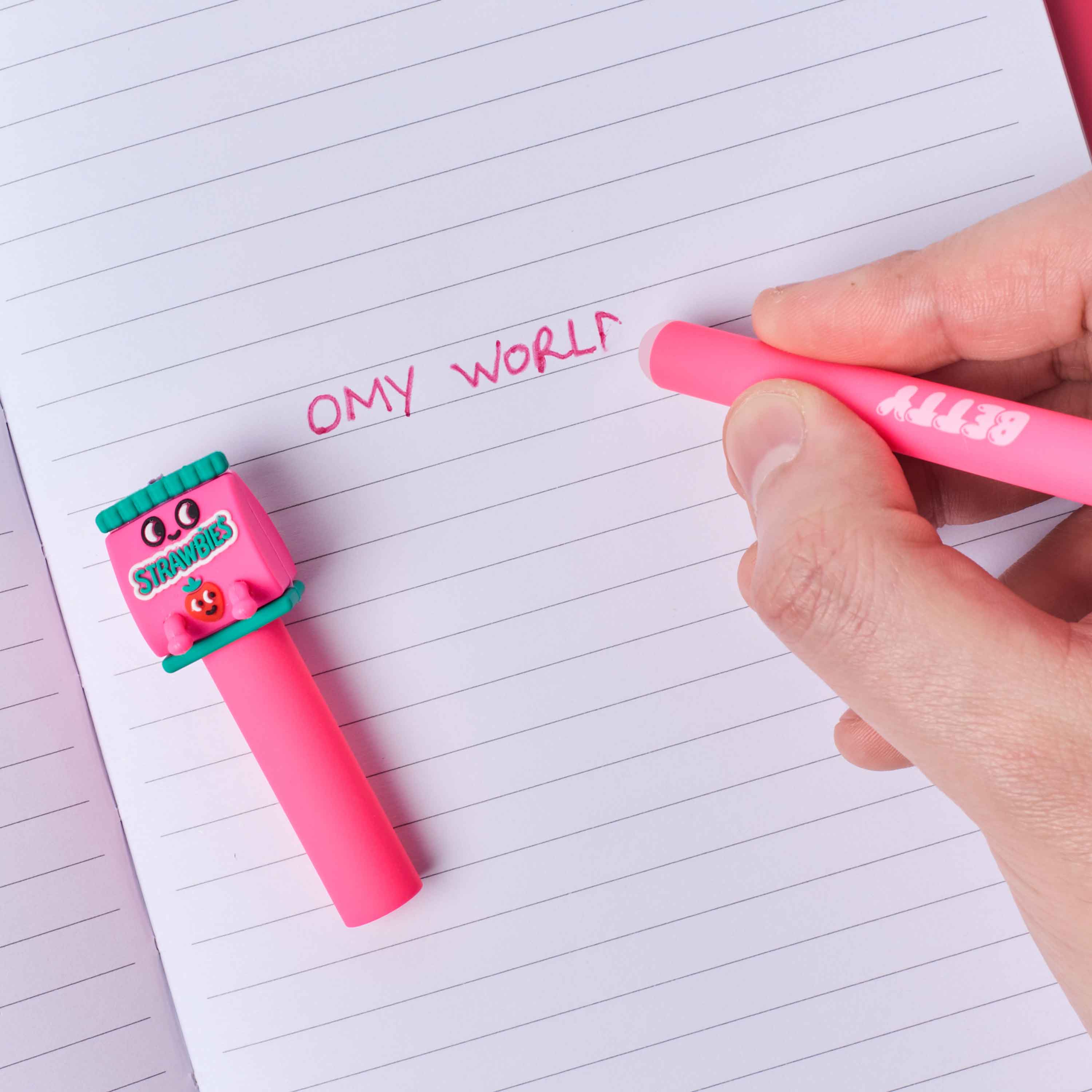 Person writing 'OMY WORLD' with a pink pen buddies betty OMY on lined paper, next to a small pink robot toy.