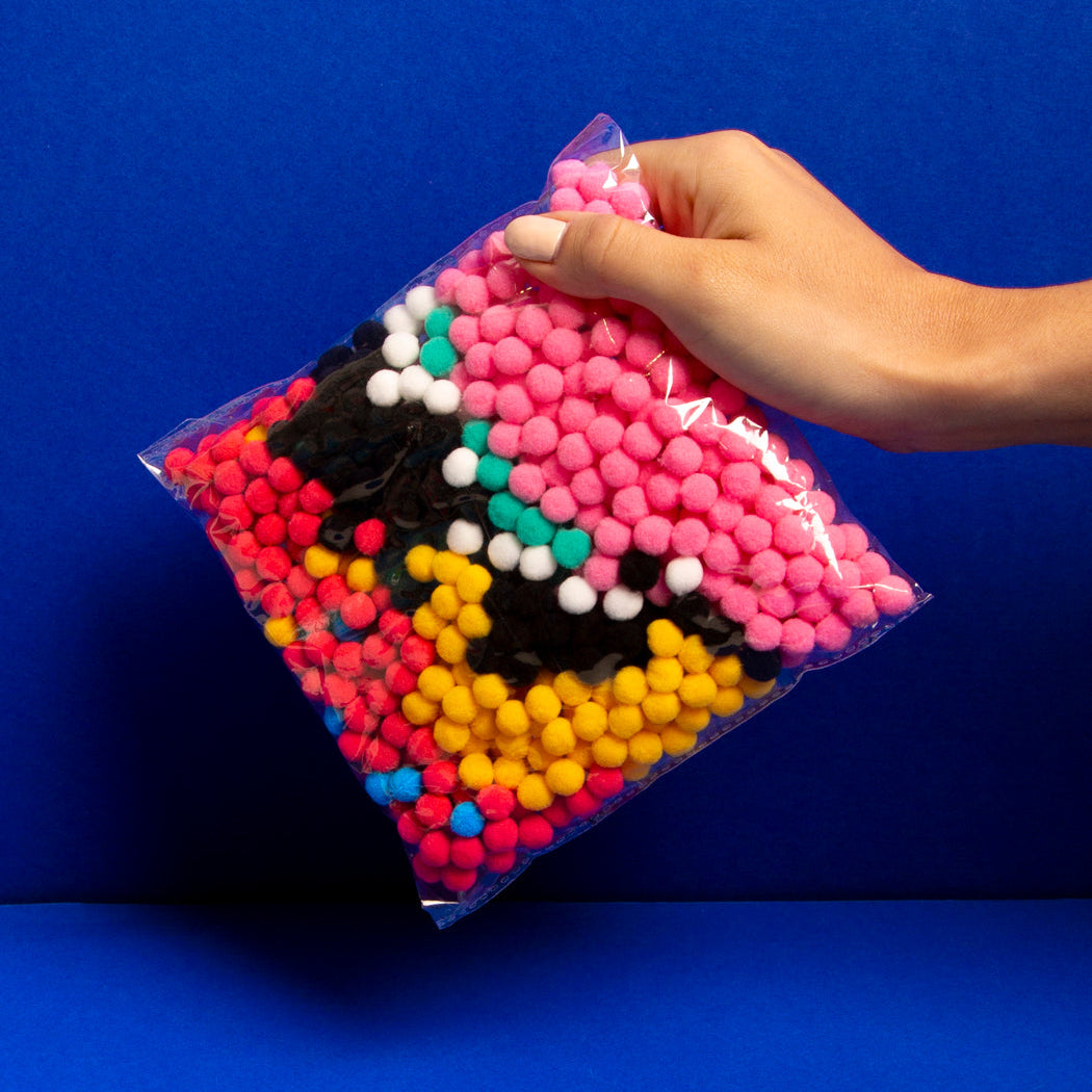 Hand holding OMY colorful pom pom craft kit forming a donut design against a blue background