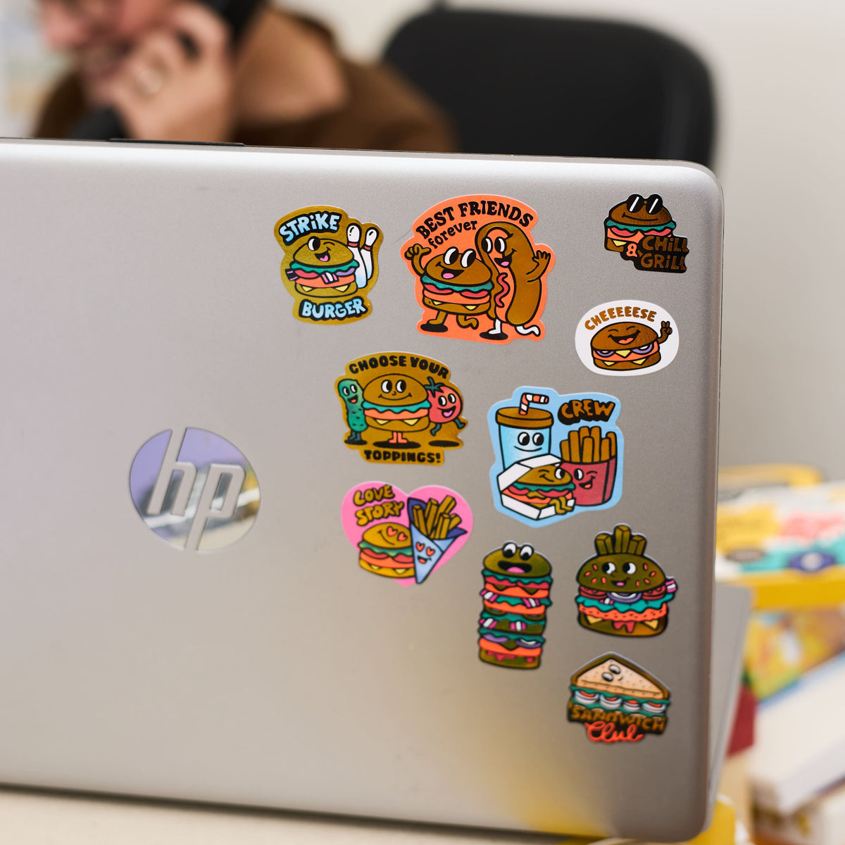 Laptop with colorful stickers big burger OMY on a desk, person using phone in the background