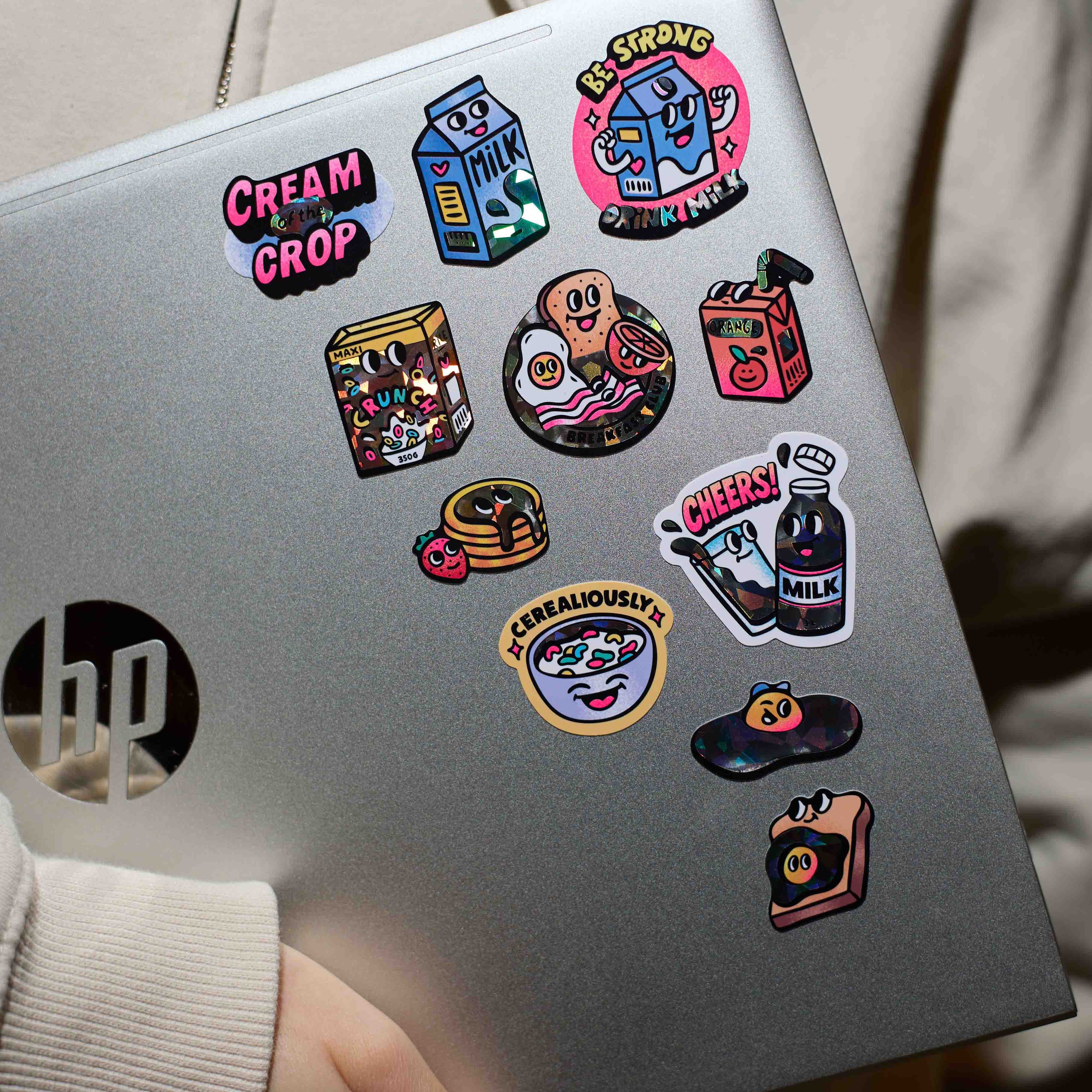 Silver HP laptop with colorful stickers milk big OMY held by a person wearing a beige jacket.