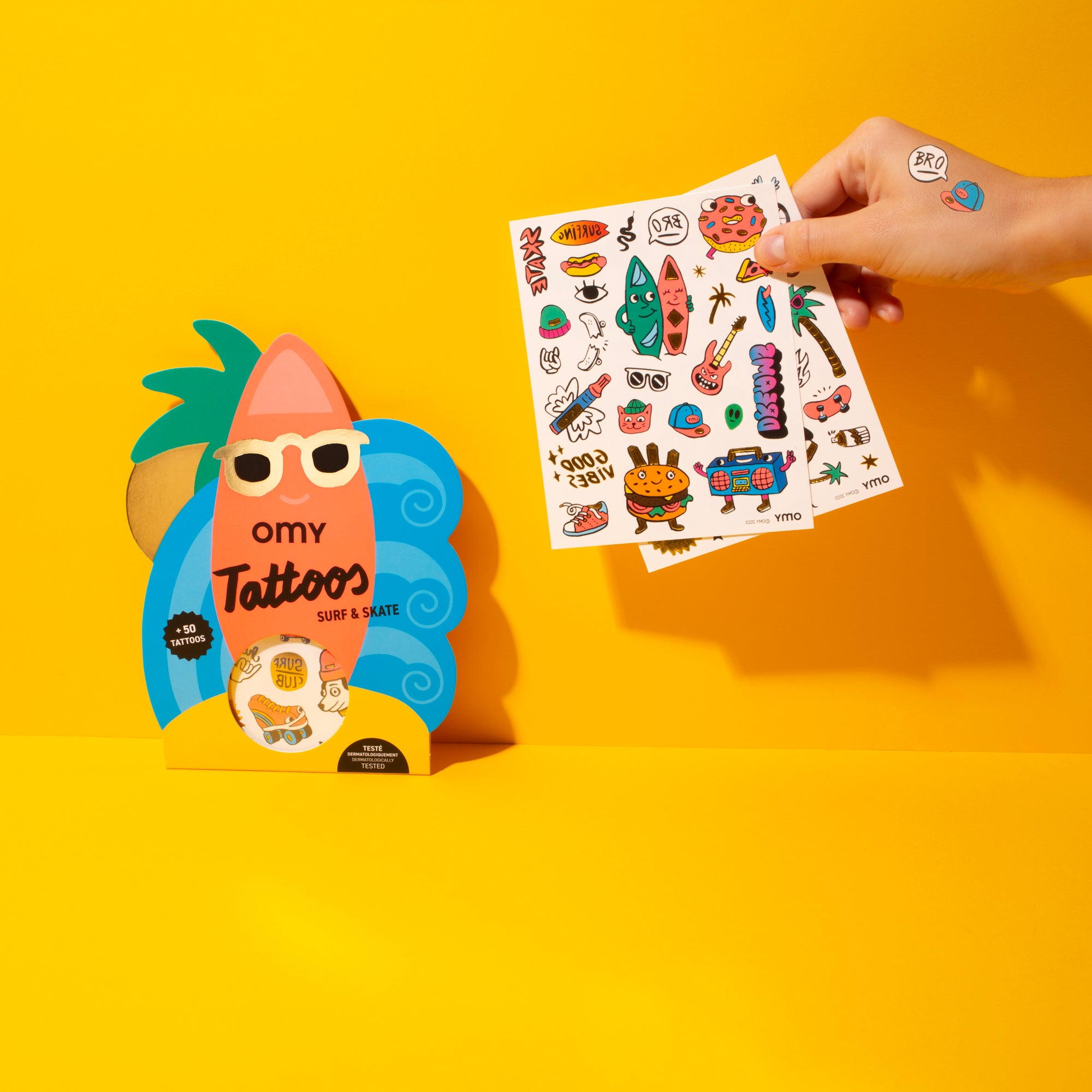 Hand holding a sheet of temporary tattoos Surf and Skate OMY with a colorful packaging on a yellow background