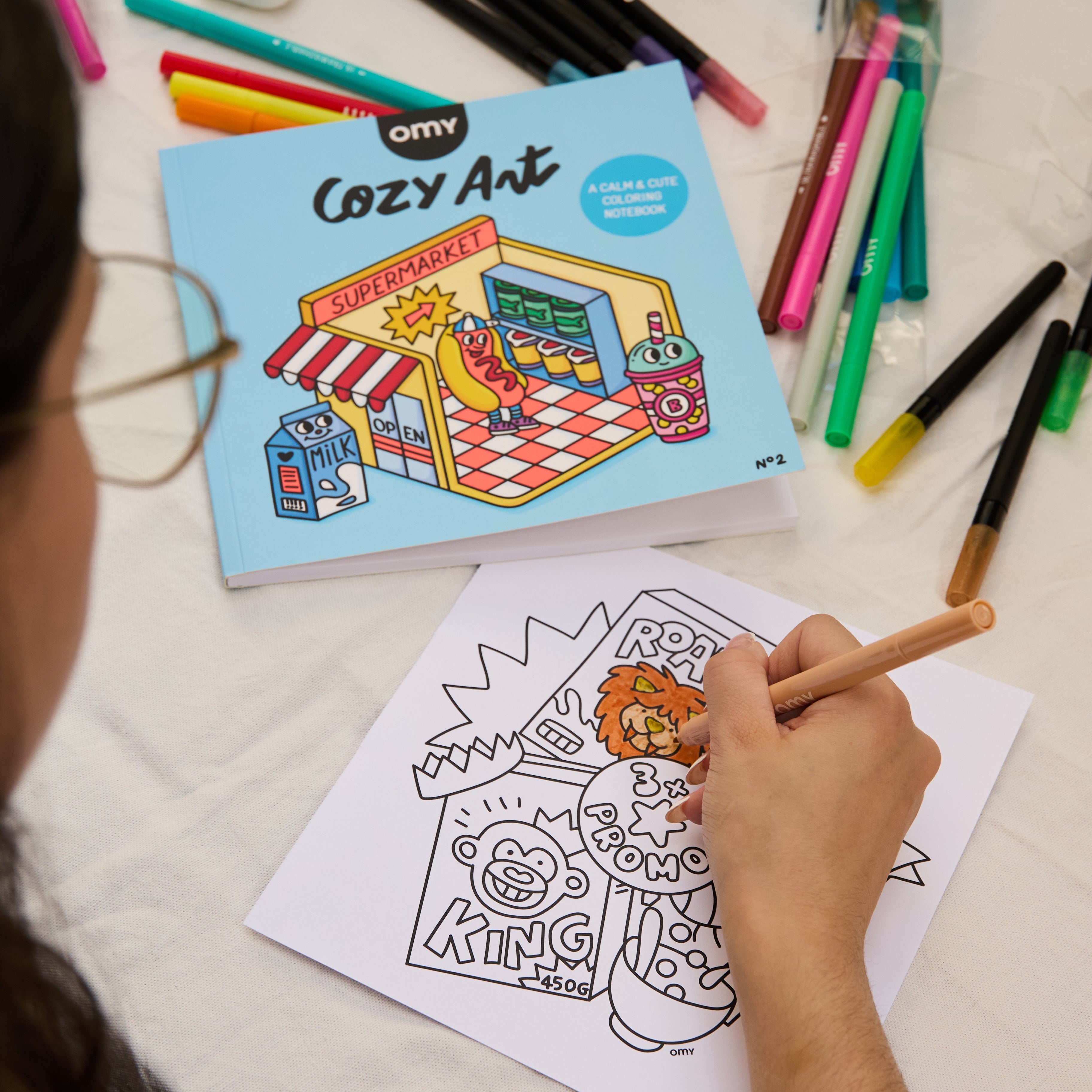Hand coloring a page from the Cozy Art "Super Market" coloring notebook, with the notebook and colorful markers in the background