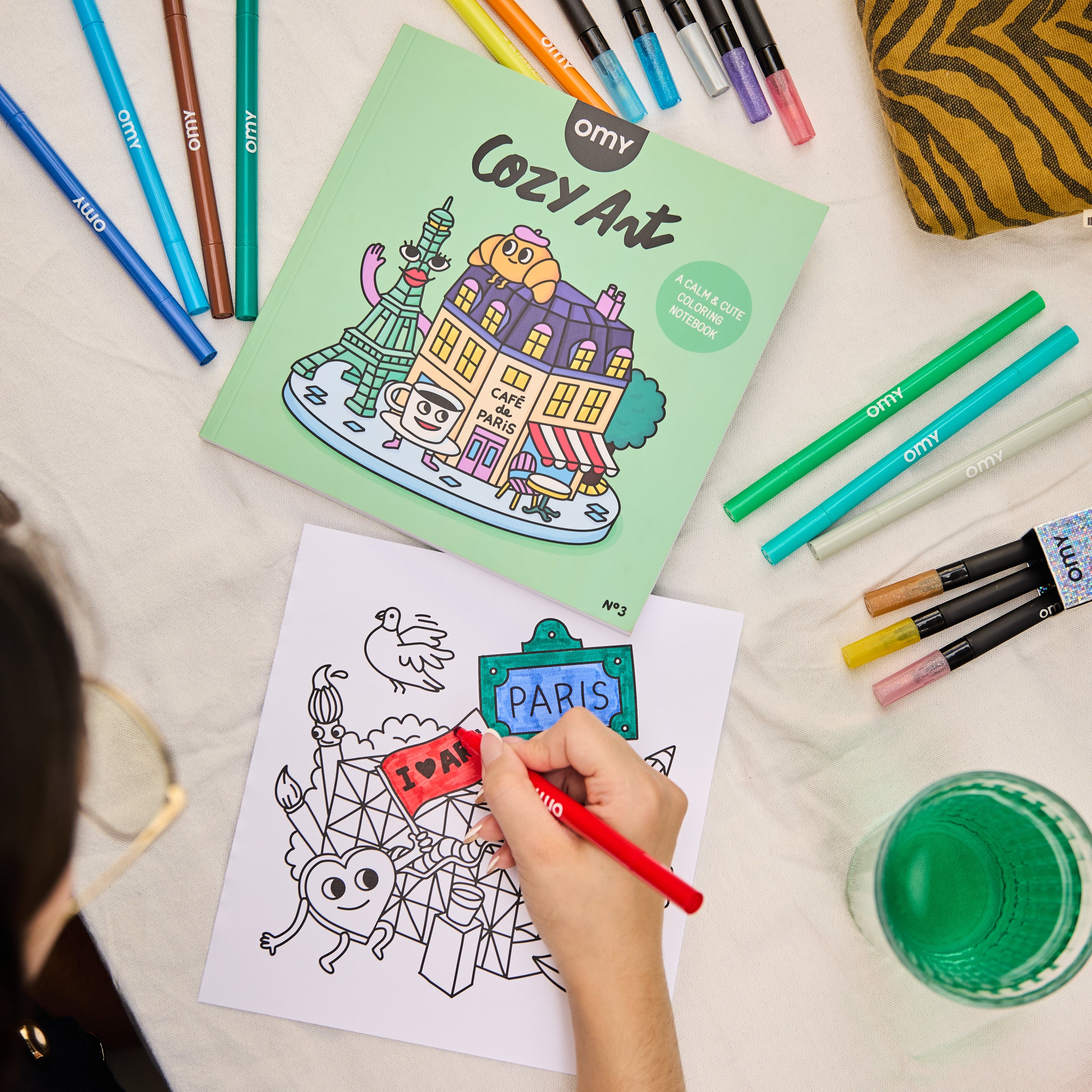 Person coloring a page from the Cozy Art Paris coloring book with vibrant markers and the book nearby, illustrating creative and relaxing coloring activity.