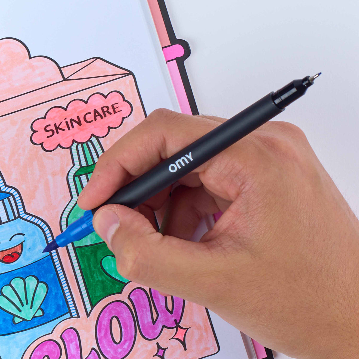 A hand holding a black Omy ballpoint pen with a blue ink tip, drawing on a colorful sketchbook page featuring cartoon skincare items and the words 'SKINCARE' and 'SLOW'.