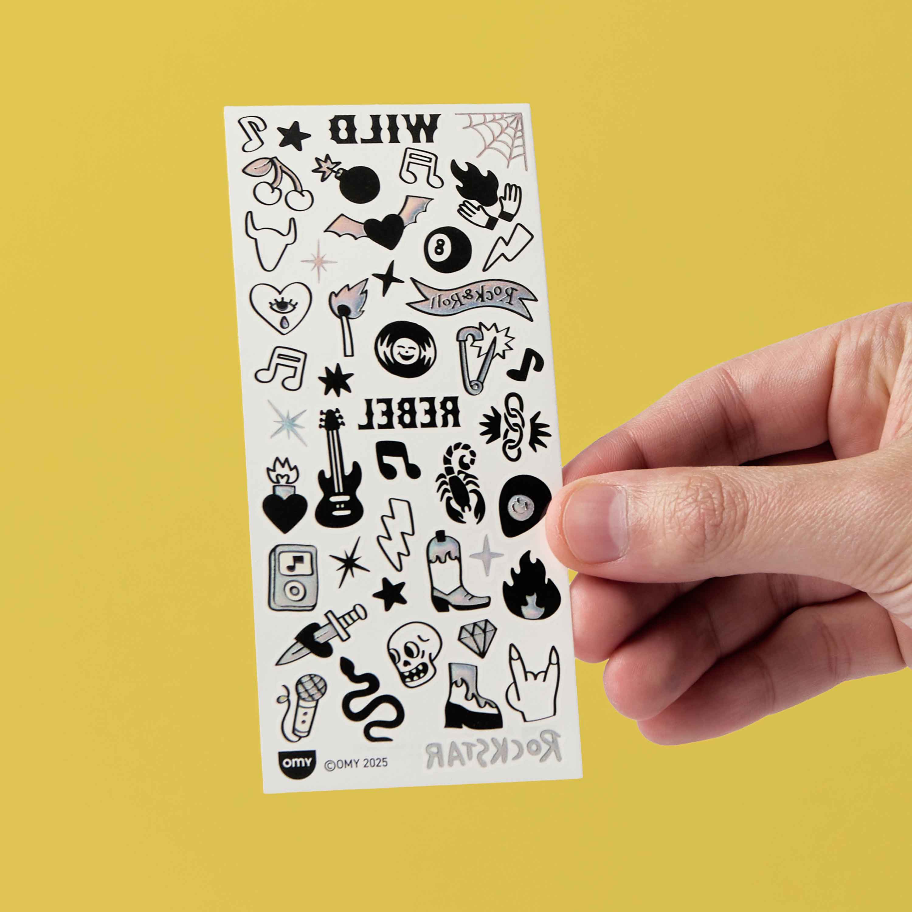 Sheet of black and white flash tattoos OMY held by a hand against a yellow background
