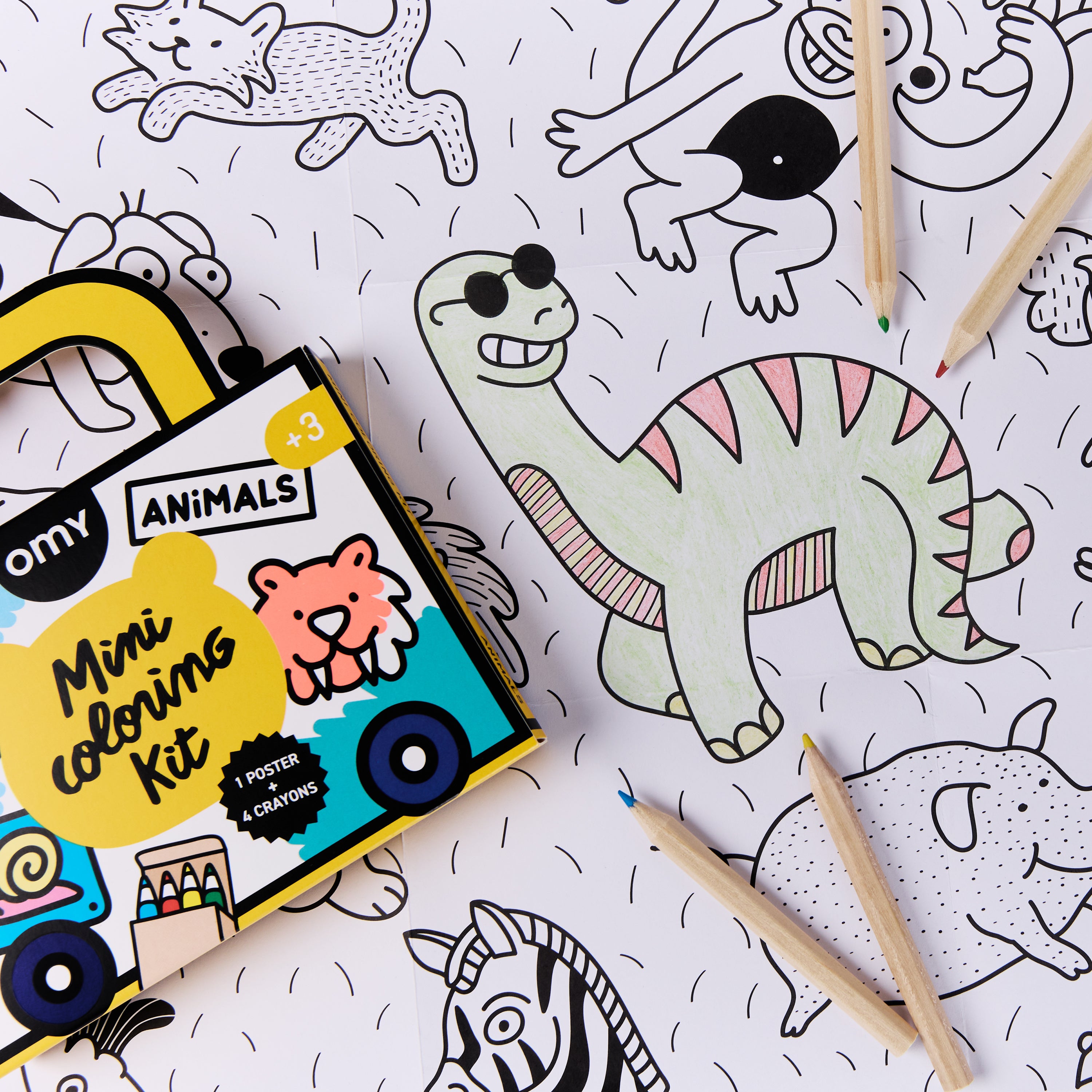 OMY Mini Coloring Kit Animals box placed on dinosaur-themed coloring poster with wooden coloring pencils; travel-sized art kit including 4 mini crayons and poster for kids ages 3 and up.