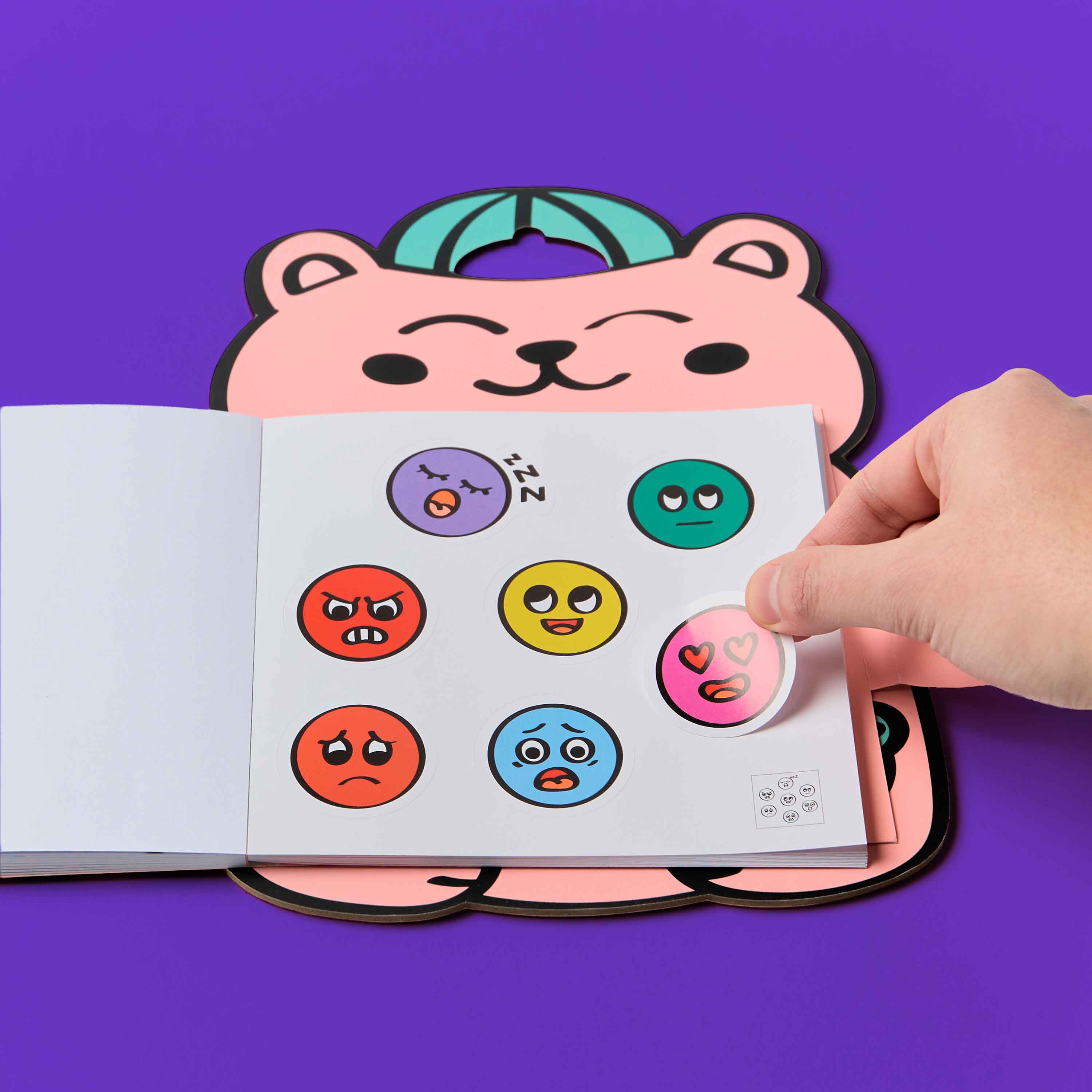 Colorful emoji stickers on a Montessorti stickers book OMY held by a hand with a pink bear-shaped object in the background.