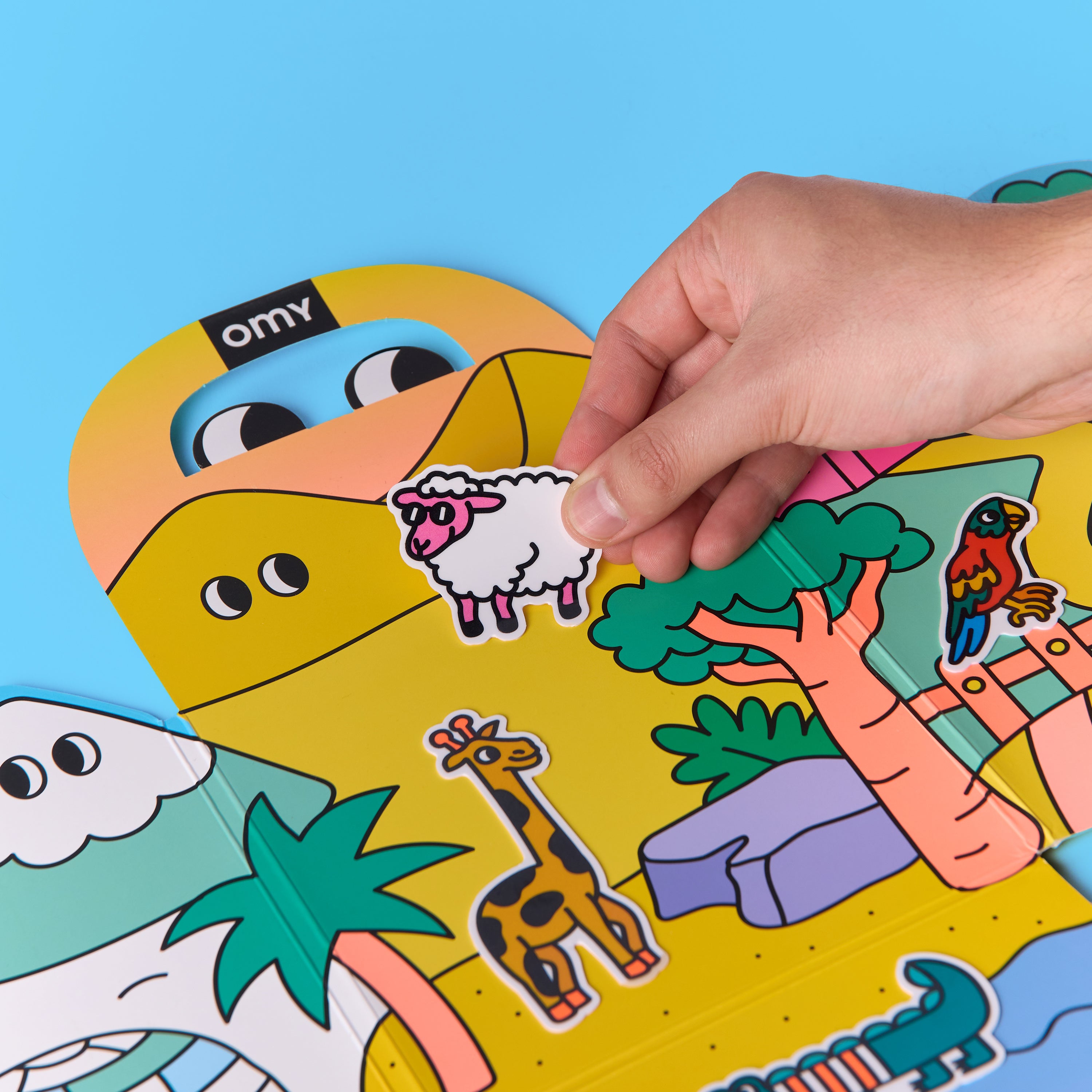 Hand placing sheep silicone sticker onto colorful Montessori animal sticker kit scene with parrot, giraffe, and trees.
