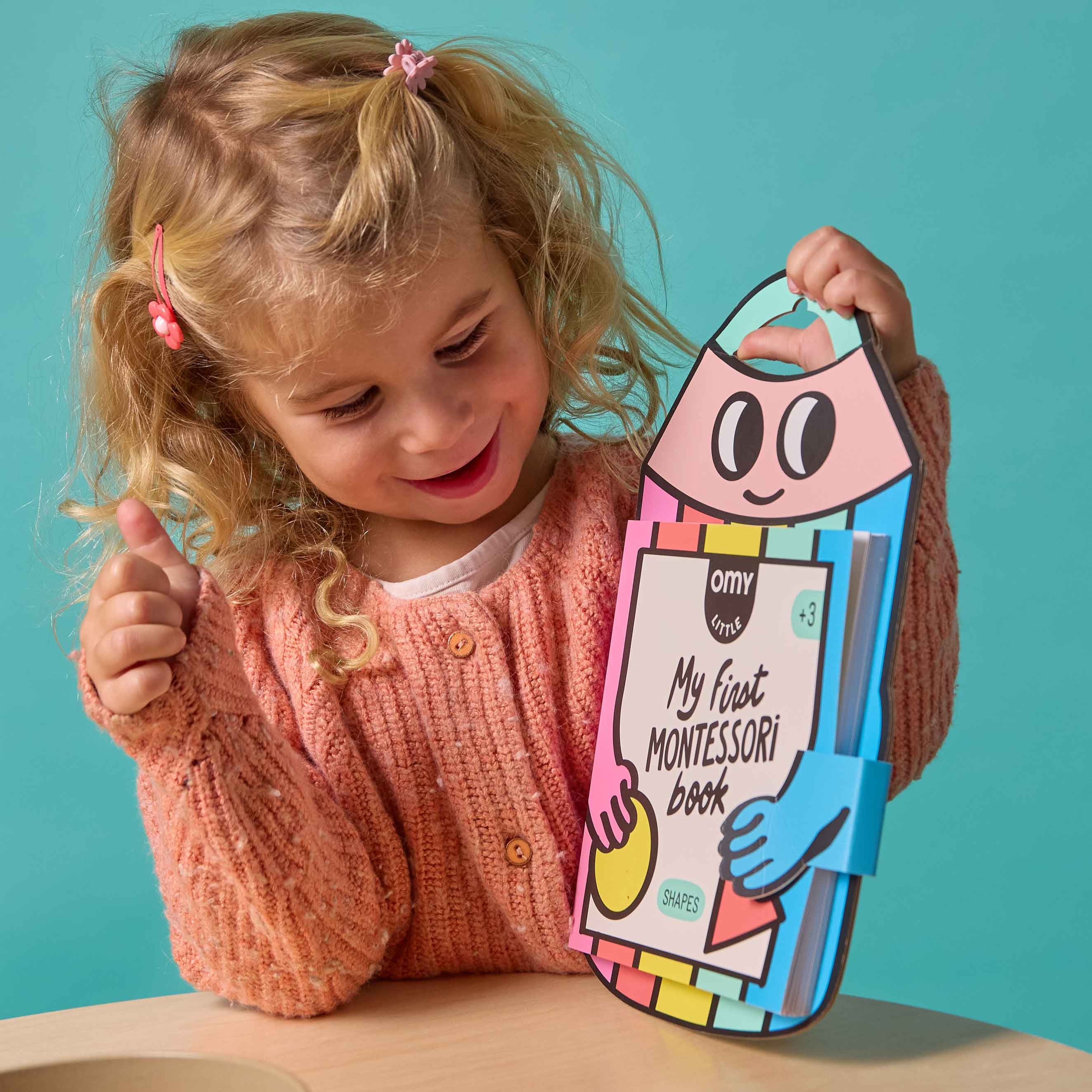 Child holding a colorful book My First Montessori Book OMY against a teal background