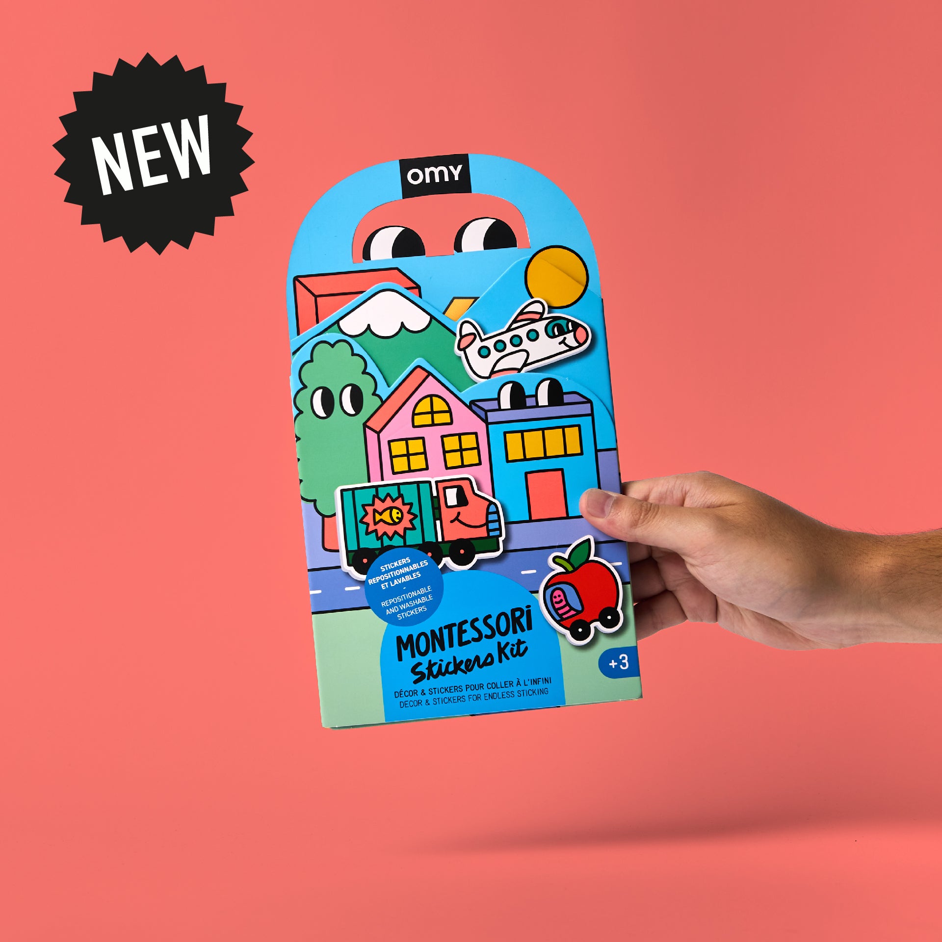 Front packaging of OMY Montessori Stickers Kit – Vroom Vroom edition featuring colorful houses, truck and helicopter on coral background with 'NEW' starburst; reusable silicone sticker kit for kids aged 3–7