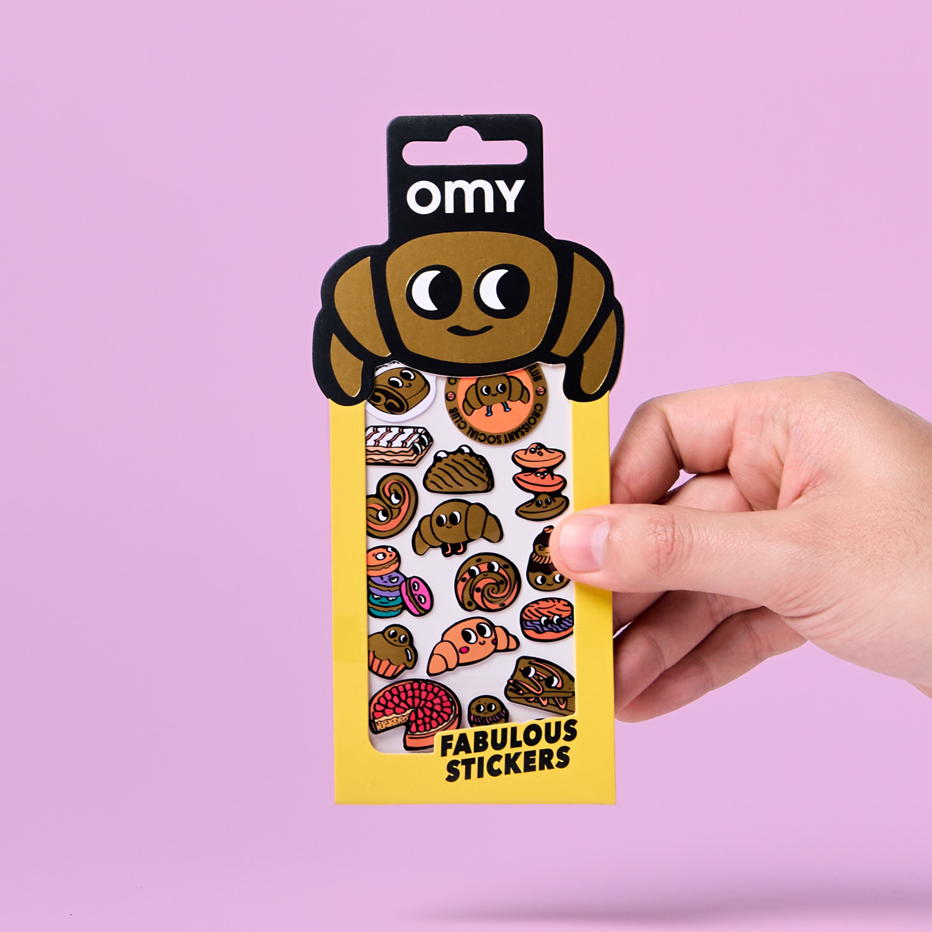 Hand holding a package of OMY stickers croissant with cartoon dog design on a pink background