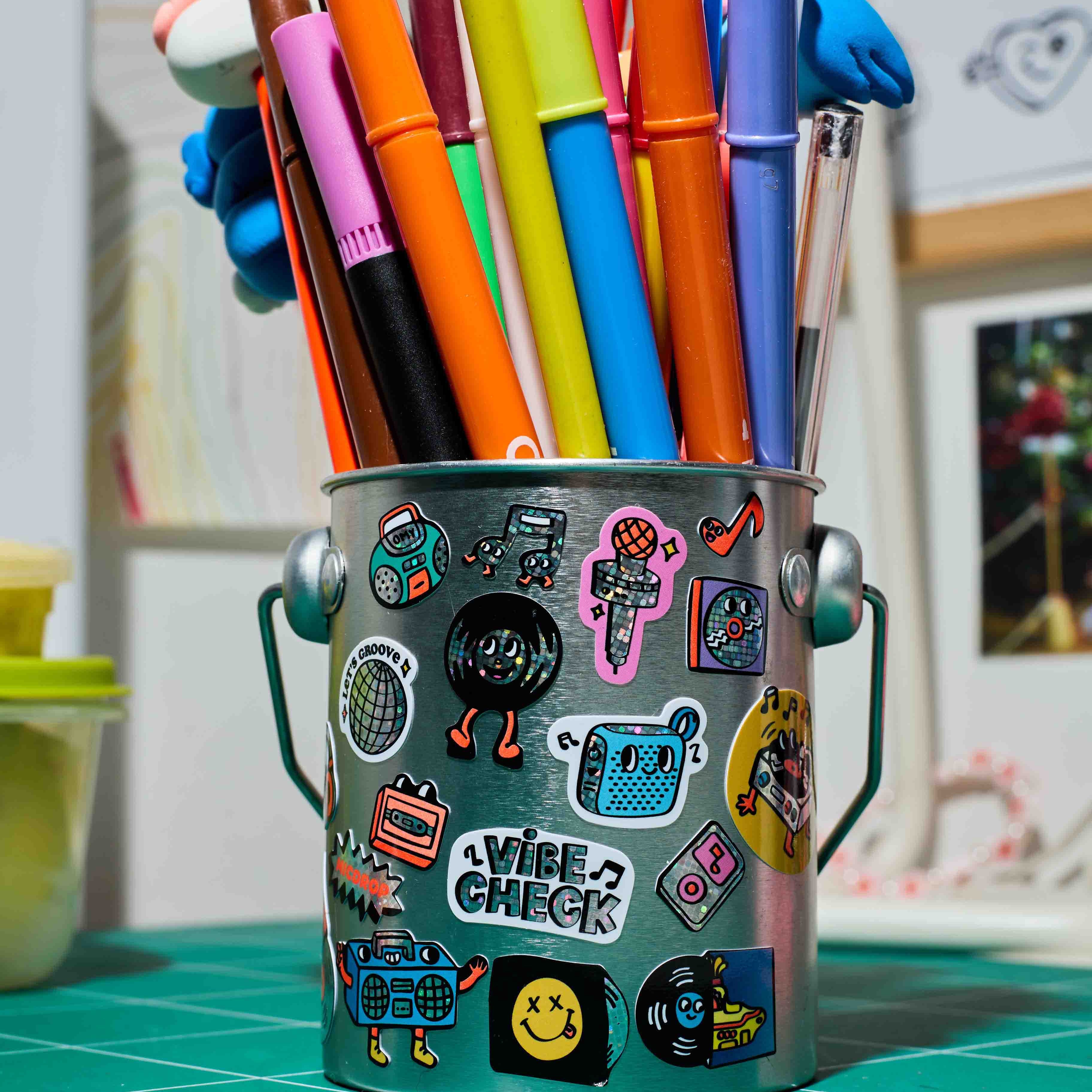 Colorful markers in a decorated metal cup  with OMY Sticker Iconic Music on a green cutting mat.